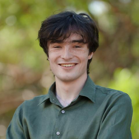 This is an image of Isaac who smiles warmly at the camera in front of a blurred park environment. He has dark hair that sits just above his brows, and is wearing an olive green collared shirt and a small silver chain.