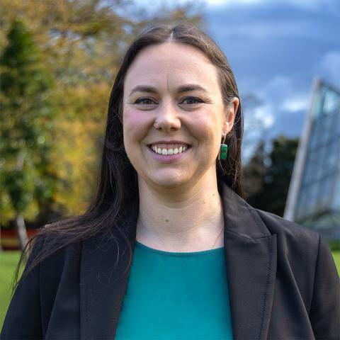 This is an image of Ellen, a warmly smiling woman in an outdoor environment. Ellen has long, dark brown hair parted in the middle with some tucked behind her ear to reveal a green stone earring. She wears a dark blazer, a green blouse and a nose ring.