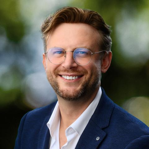 This is an image of Angel who is smiling warmly at the camera. He has short, light brown hair that frames his face and neat, stubbly beard. He's wearing a navy jacket on top of a collared white shirt with round, silver-rimmed glasses.