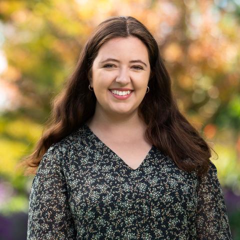 This is an image of Angelica who is smiling at the camera in an outdoor environment. She has long, dark, wavy hair, is wearing a black printed blouse and small, golden hoop earrings. The background includes vibrant foliage in autumnal colours and looks bright and warm.