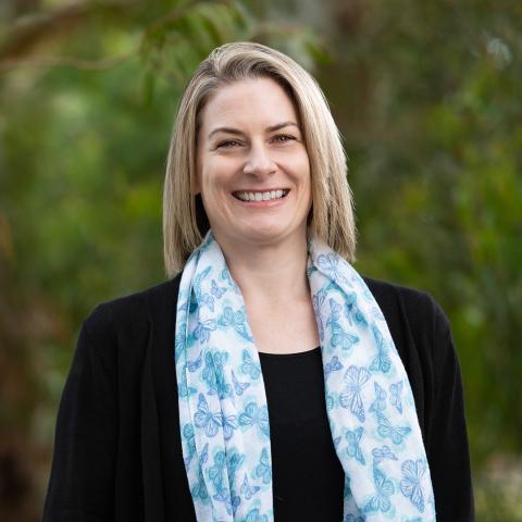 Profile picture for Dr Merran Blair, Greens candidate for Monbulk. Merran is standing in front of blurred out trees and is wearing a black top with a light blue scarf with butterflies on it