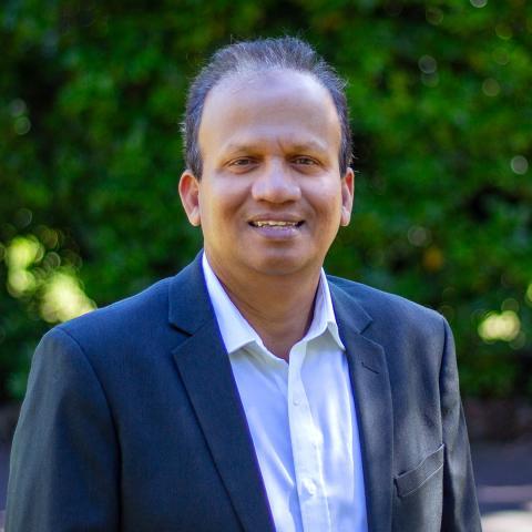 A man named Rifai, wearing glasses and a navy blazer over a white collared shirt, stands smiling against a backdrop of lush green foliage. The natural setting and his relaxed demeanor convey warmth and professionalism.