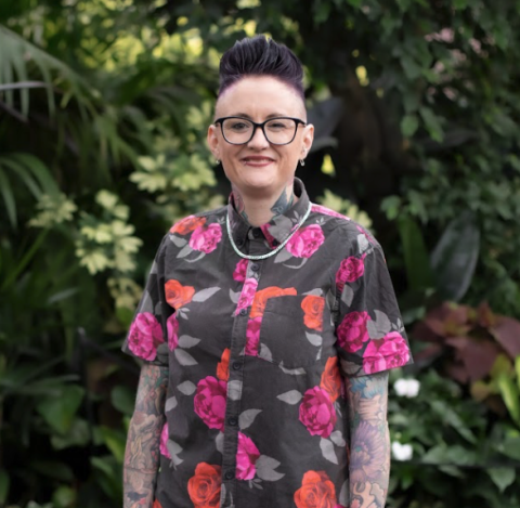 This is an image of Jess, a smiling individual with glasses, short hair and a black button up shirt that has red and pink roses on it. Jess is standing outside, in front of a dark green tree and shrubbery background.