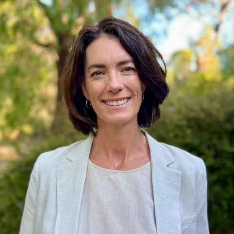 Sianan smiles at the camera, stood in front of a green, park environment. She has dark brown hair, that sits above her shoulders and is tucked back to reveal small hooped earrings. She wears a light coloured blazer and shirt.