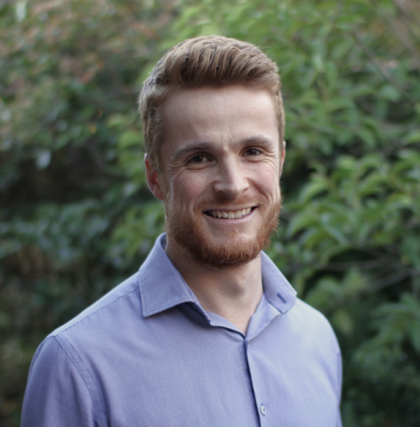 This is an image of a man named Michael, standing outdoors, wearing a blue collared shirt and smiling in front of a dark green, leafy background. 