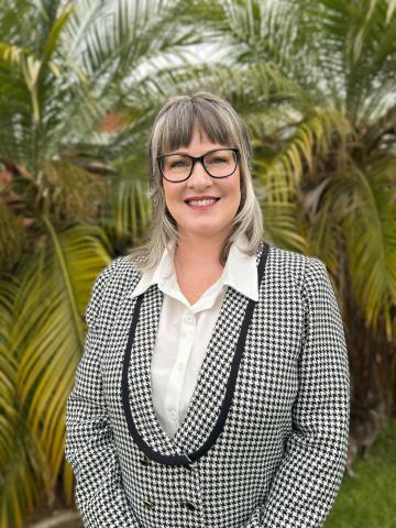 A woman with shoulder-length hair, wearing a stripy blazer and white button up shirt. Standing in front of a tree, smiling.