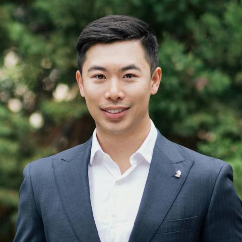 This is an image of Aaron, who is smiling warmly at the camera in front of a dark green, outdoor environment. He has short dark hair, and wears a white collared shirt under a great suit jacket.