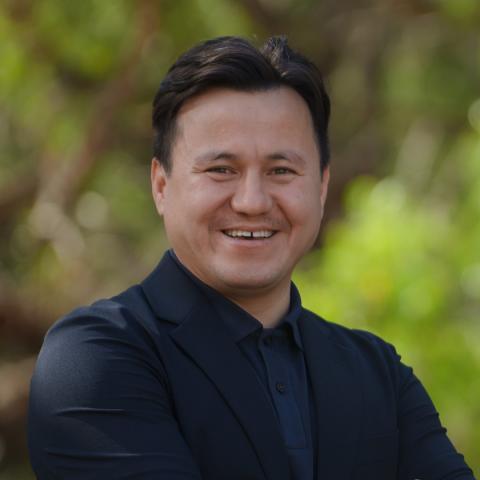 This is an image of Nasser, who smiles brightly at the camera in front of a blurred outdoor environment. He wears a black collared shirt and jacket, with neatly cut, short dark hair.