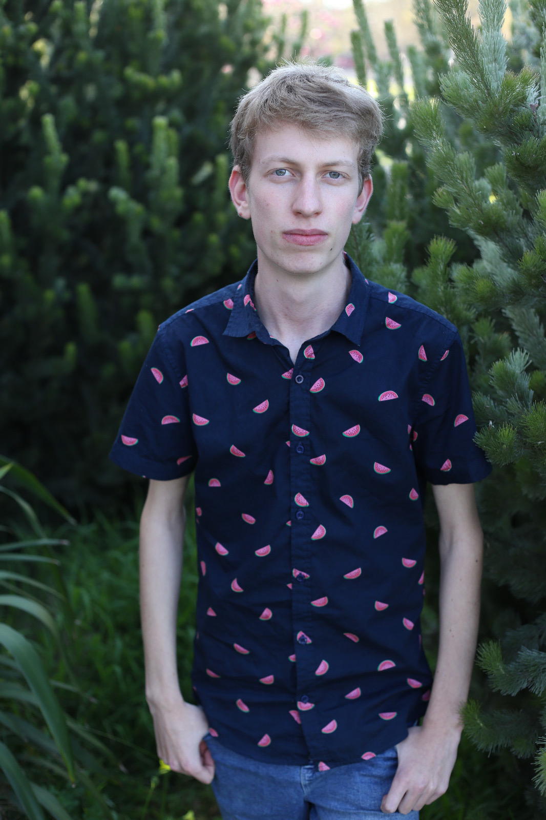 Owen is standing outdoors, wearing a navy blue shirt with pink watermelon patterns. He has short, light brown hair and a neutral expression. The background is filled with green foliage and pine trees.