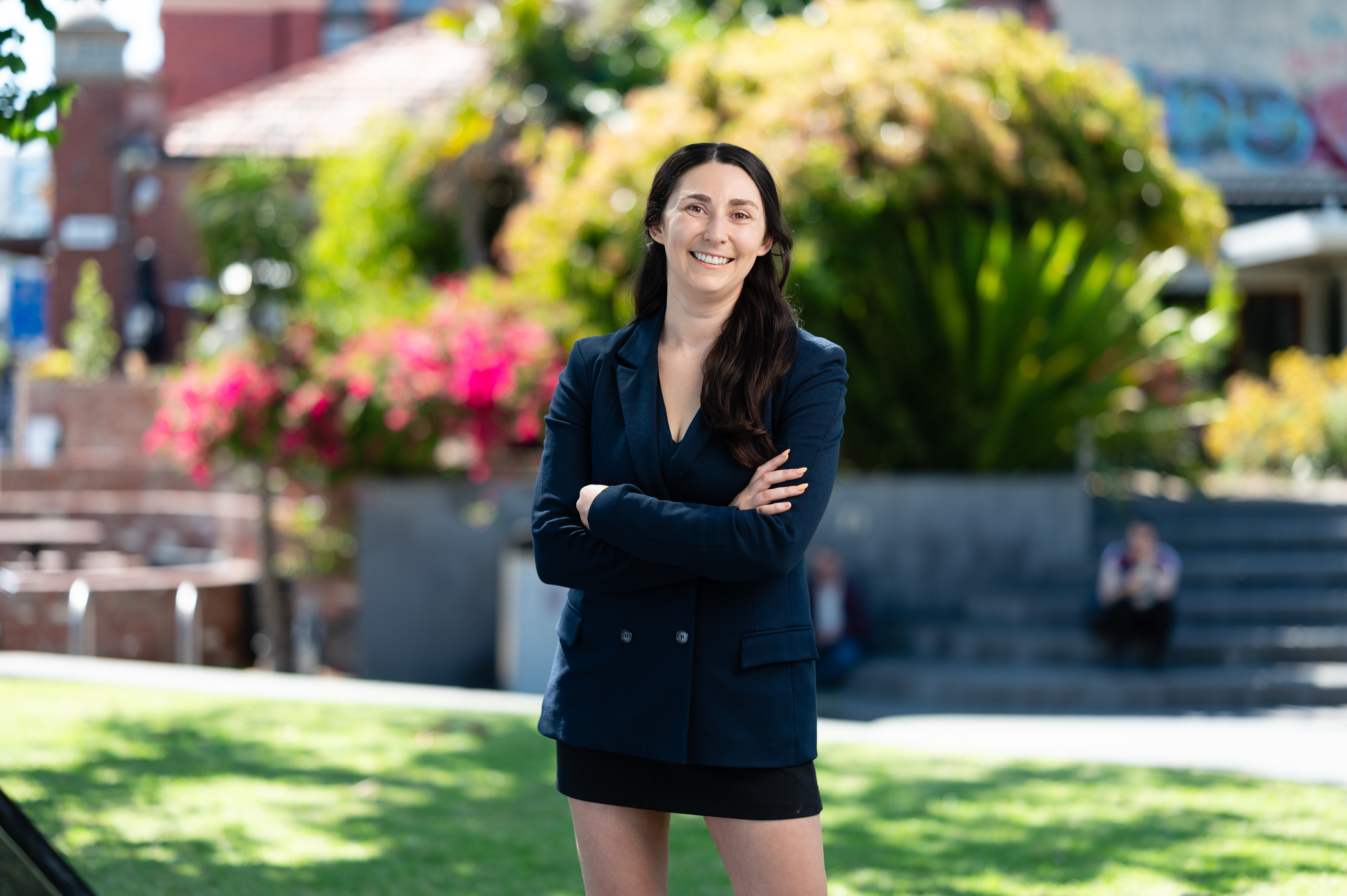 Alana stands outdoors in a navy blazer and black skirt, smiling confidently with arms crossed. She has long dark hair, and the background features vibrant greenery, flowers, and urban elements under bright sunlight.