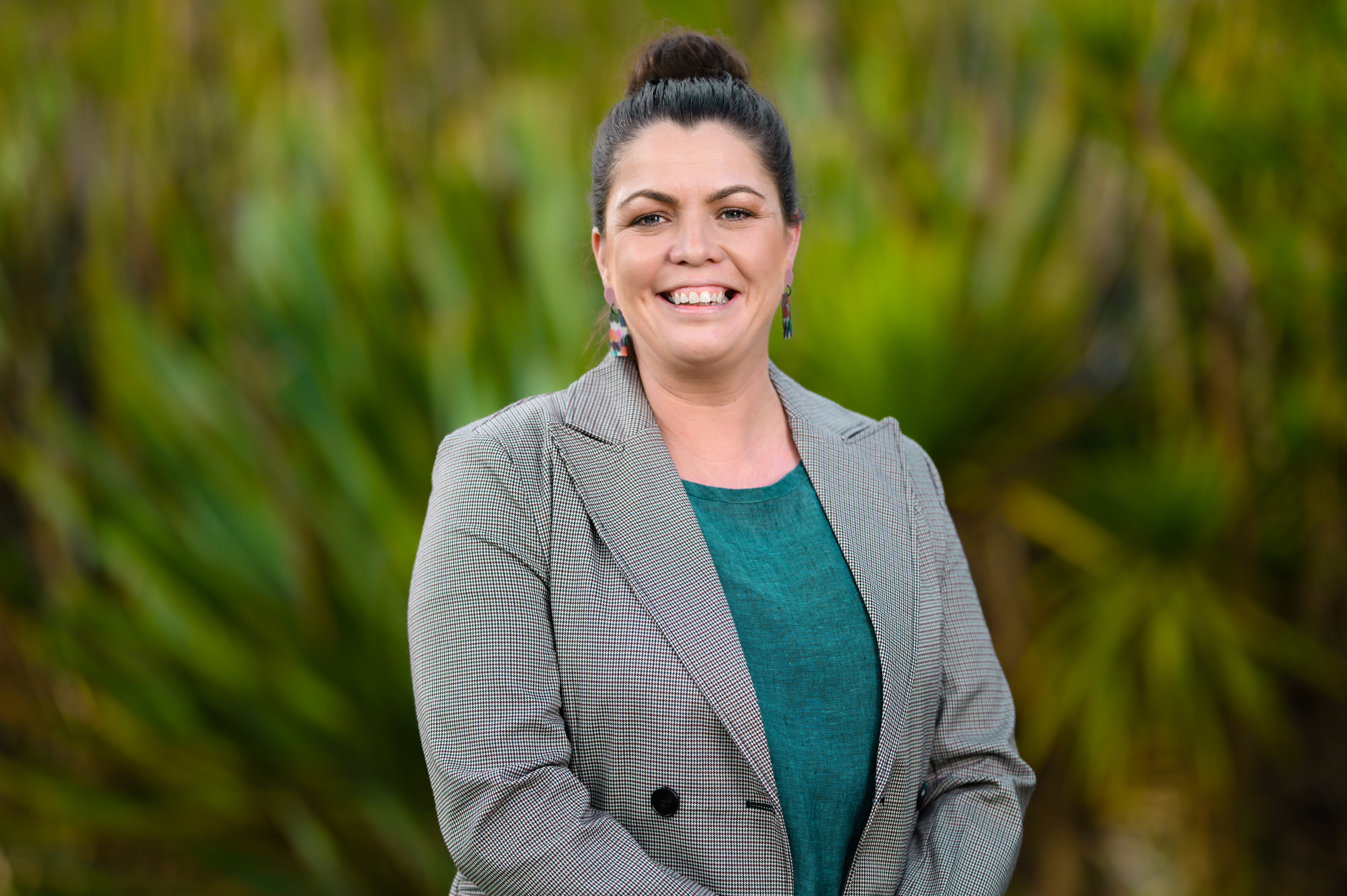 Alysia Regan, a woman with dark hair in a bun, smiles warmly. She wears a green top, a checkered blazer, and colorful earrings. The background is blurred greenery, suggesting an outdoor setting.