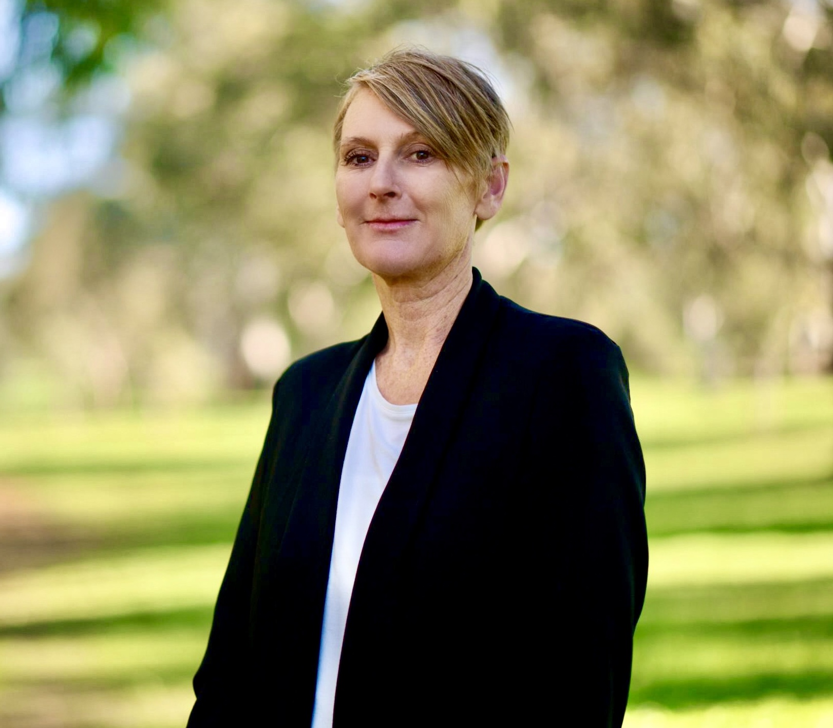 A woman with short blonde hair, a black blazer and white shirt in a leafy park.