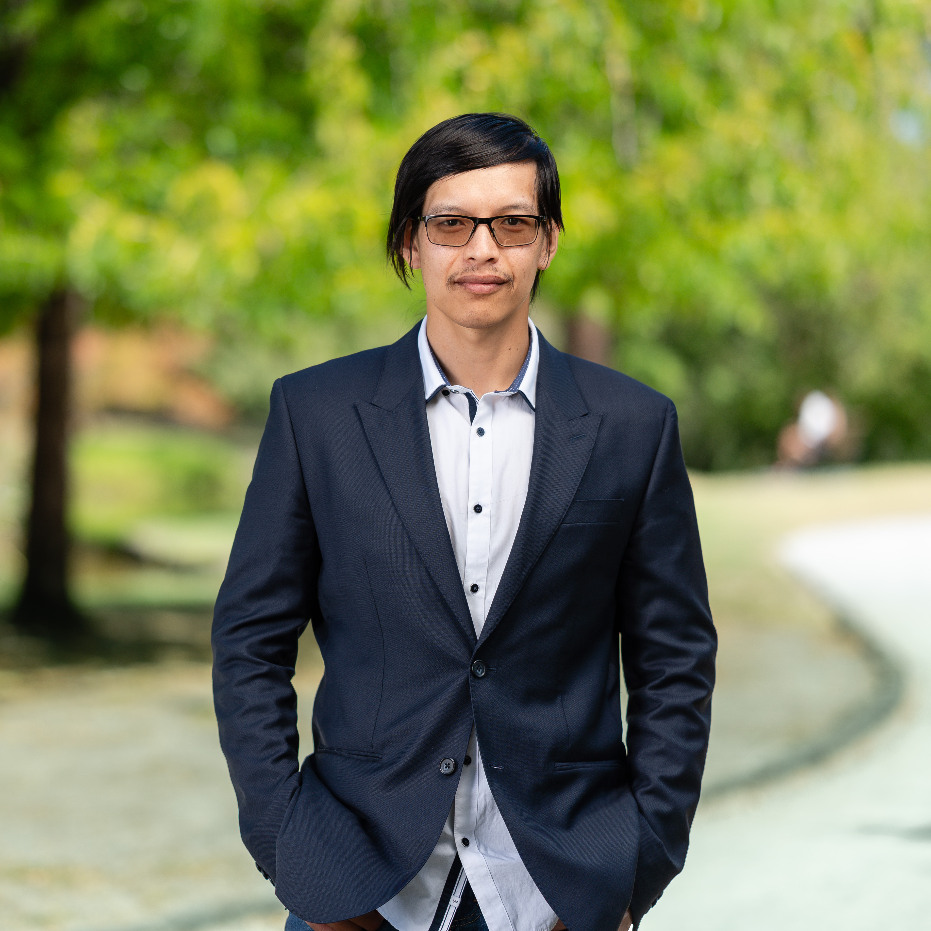 Thuc Bao Huynh, a man with black hair and glasses, stands outdoors wearing a navy blazer over a white shirt. He has a calm expression with hands in pockets. The background is a green park with trees and a winding path.