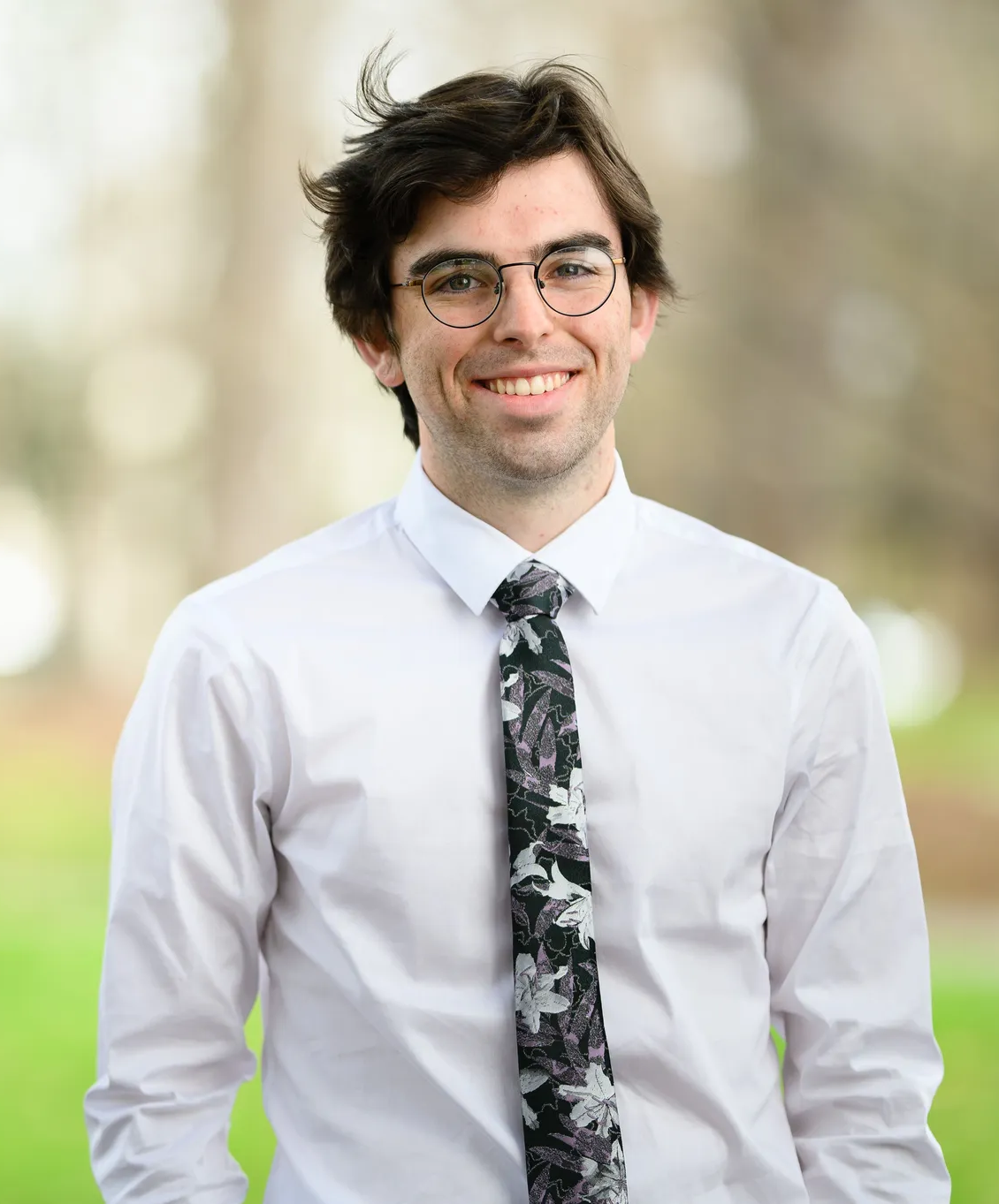 Callum wears glasses, a white shirt and black patterned tie. He is standing in front of blurred green foliage.