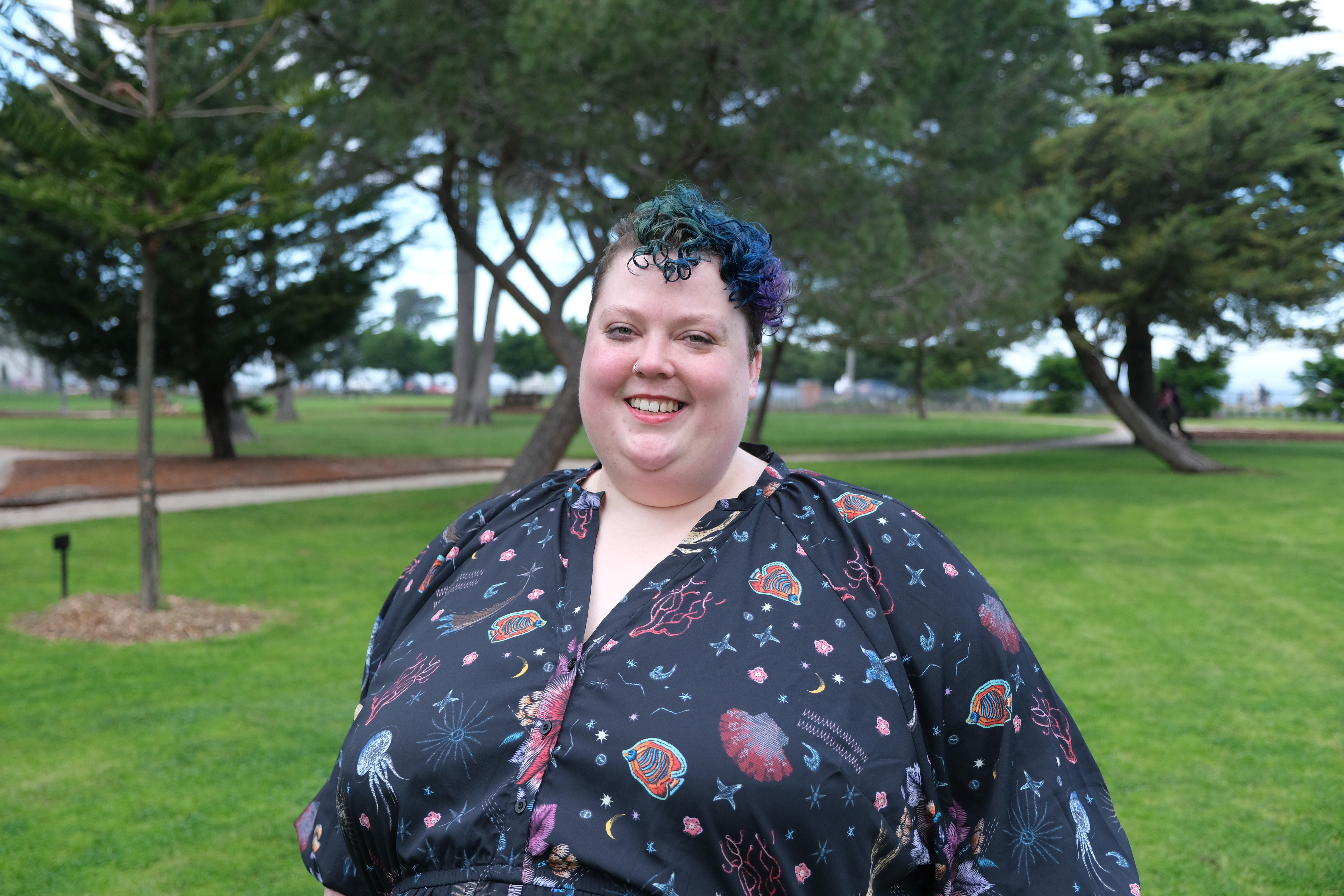 Maggie Ralph standing in front of trees with blue and purple hair smiling