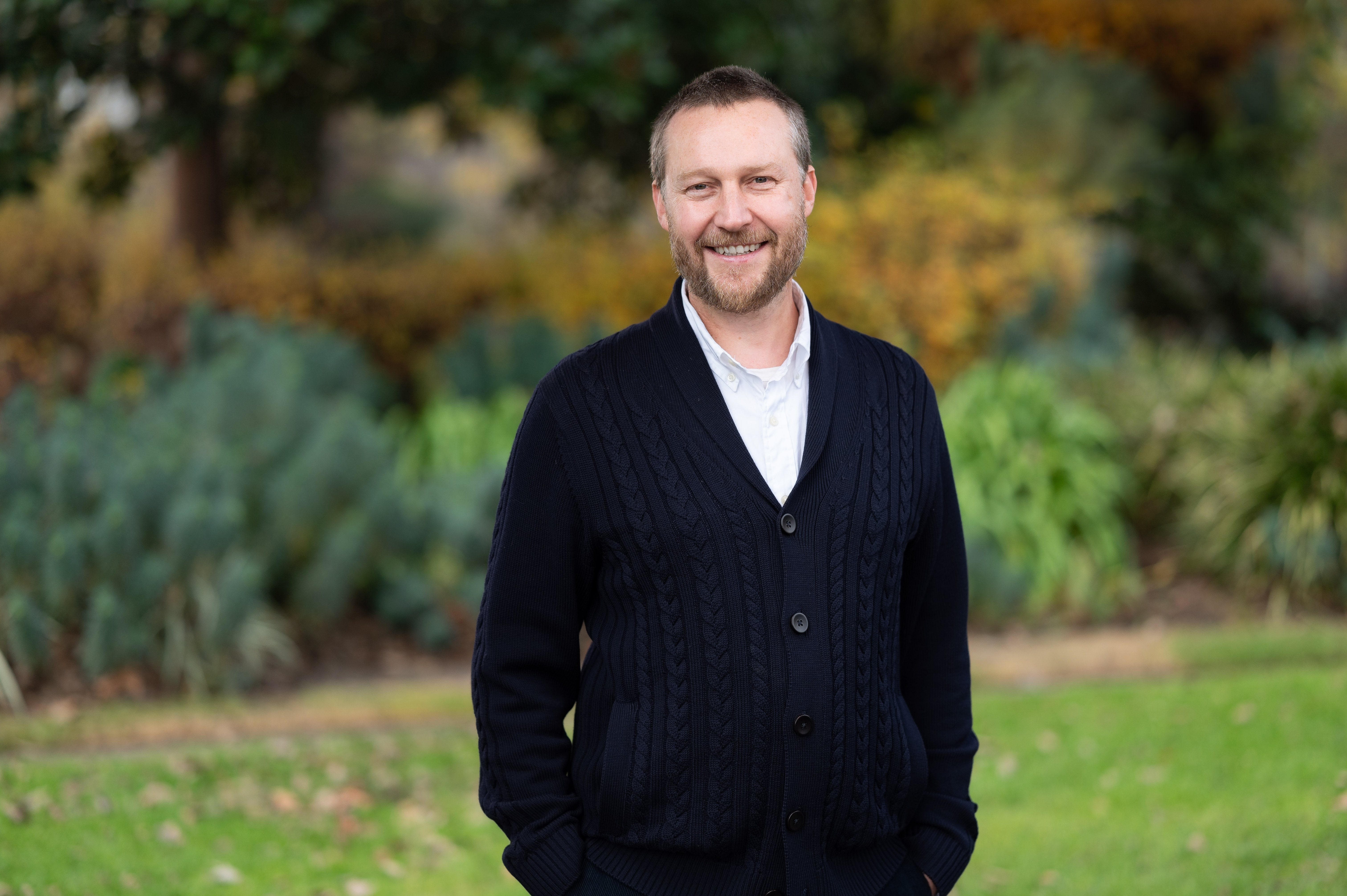 A man named Dave Perry stands outdoors, smiling, wearing a navy blue cardigan over a white shirt. The background shows blurred greenery and foliage.