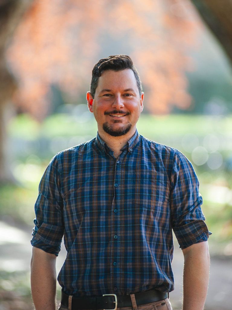 Clint Uink standing in a leafy park, wearing a checked shirt, smiling at camera.