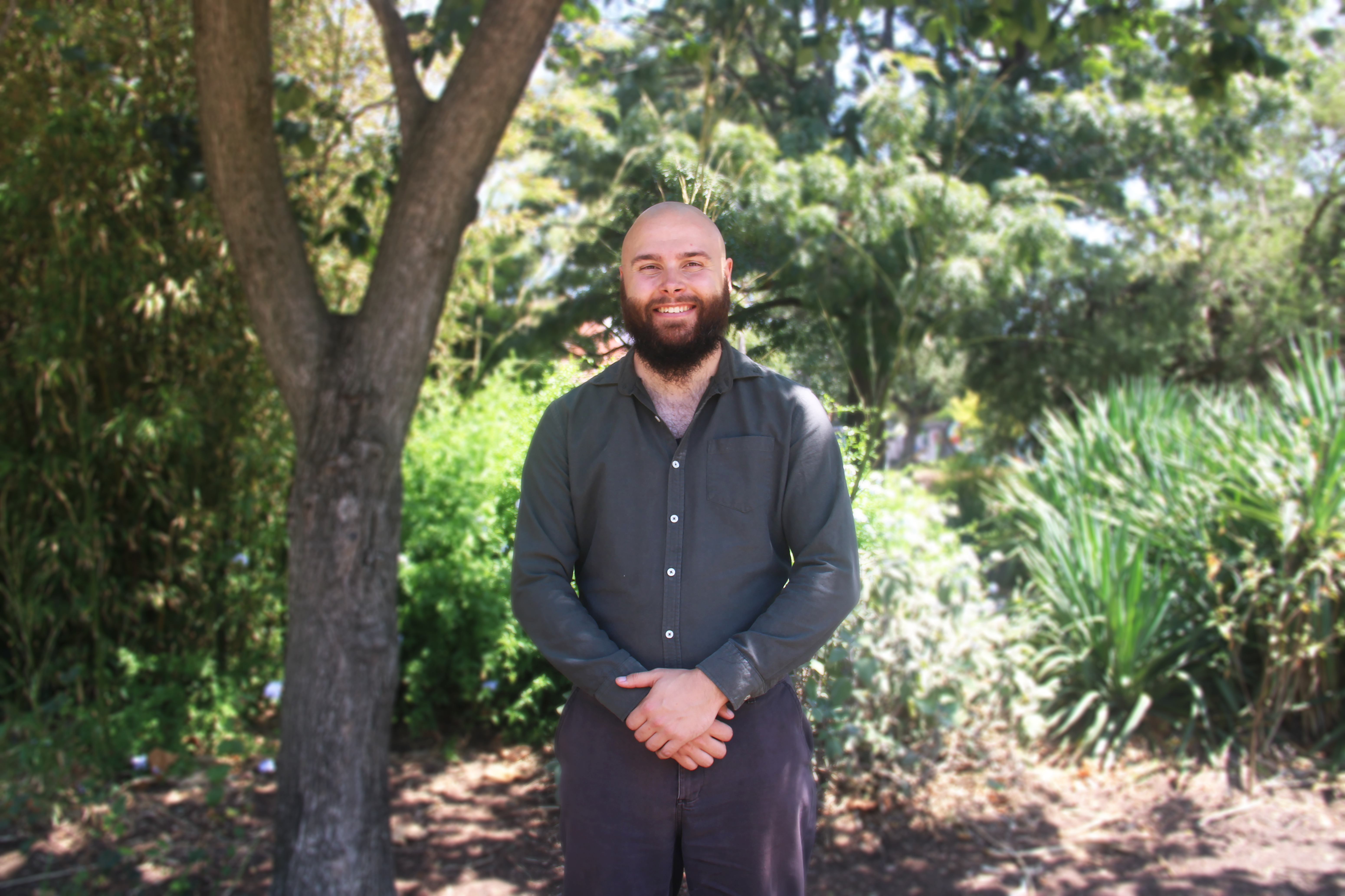 Matt Maber stands under a tree, smiling warmly. He wears a dark button-up shirt and dark pants. Sunlight filters through the leaves, casting dappled light on the lush greenery behind him.