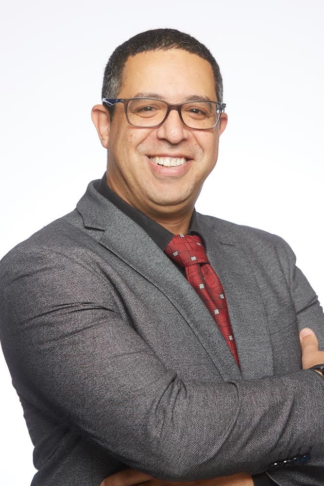 A man named Mohamed El-Mastri, wearing glasses, a gray suit, and a red tie with a subtle pattern, smiles with his arms crossed. He stands against a plain white background.