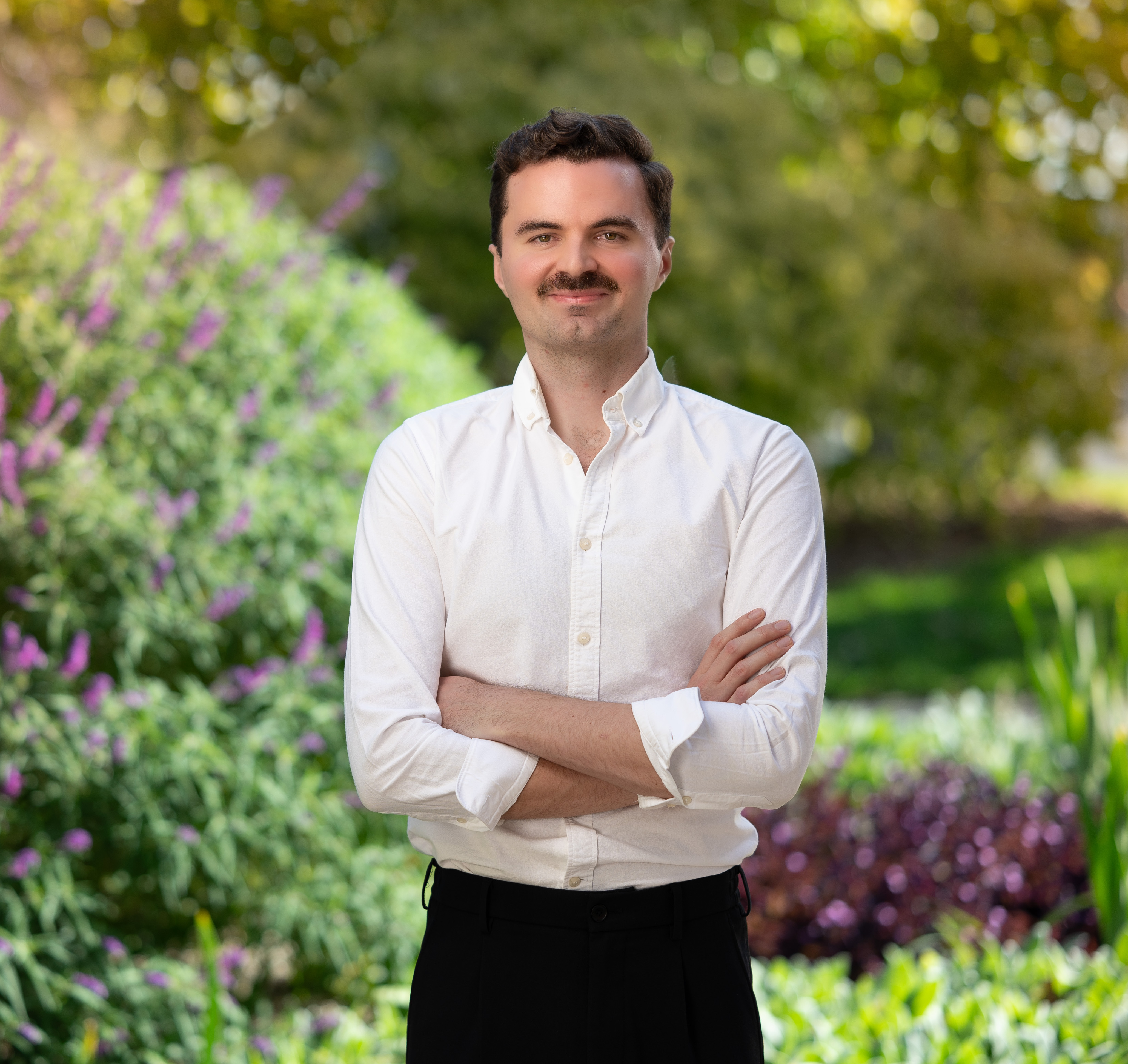 A man with a white button up shirt, black pants and a black moustache. Standing with his arms crossed and smiling in front of a leafy background.