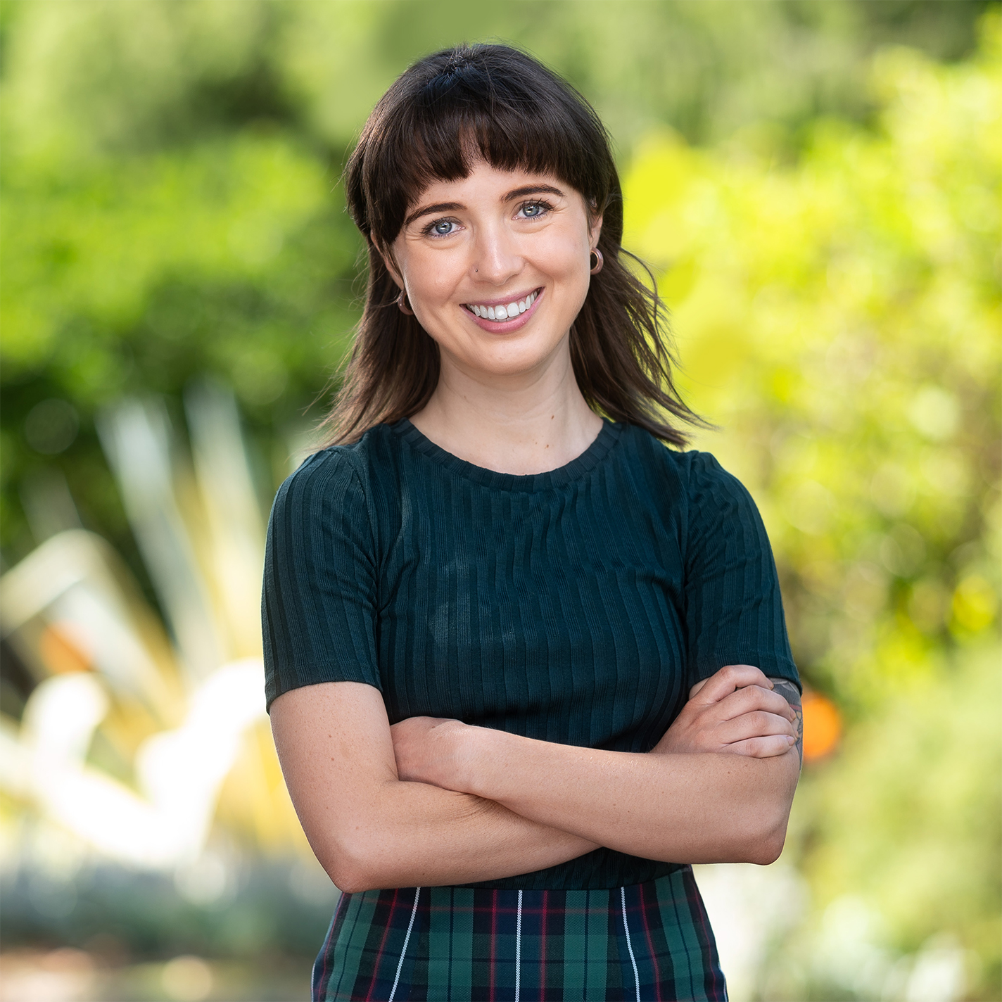 This is an image of Amy, a smiling individual standing outdoors in a lush natural environment. She has short dark hair with bangs, is wearing a fitted green shirt, and checked skirt. The background includes vibrant green foliage and tall grasses creating a calm and pleasant setting.