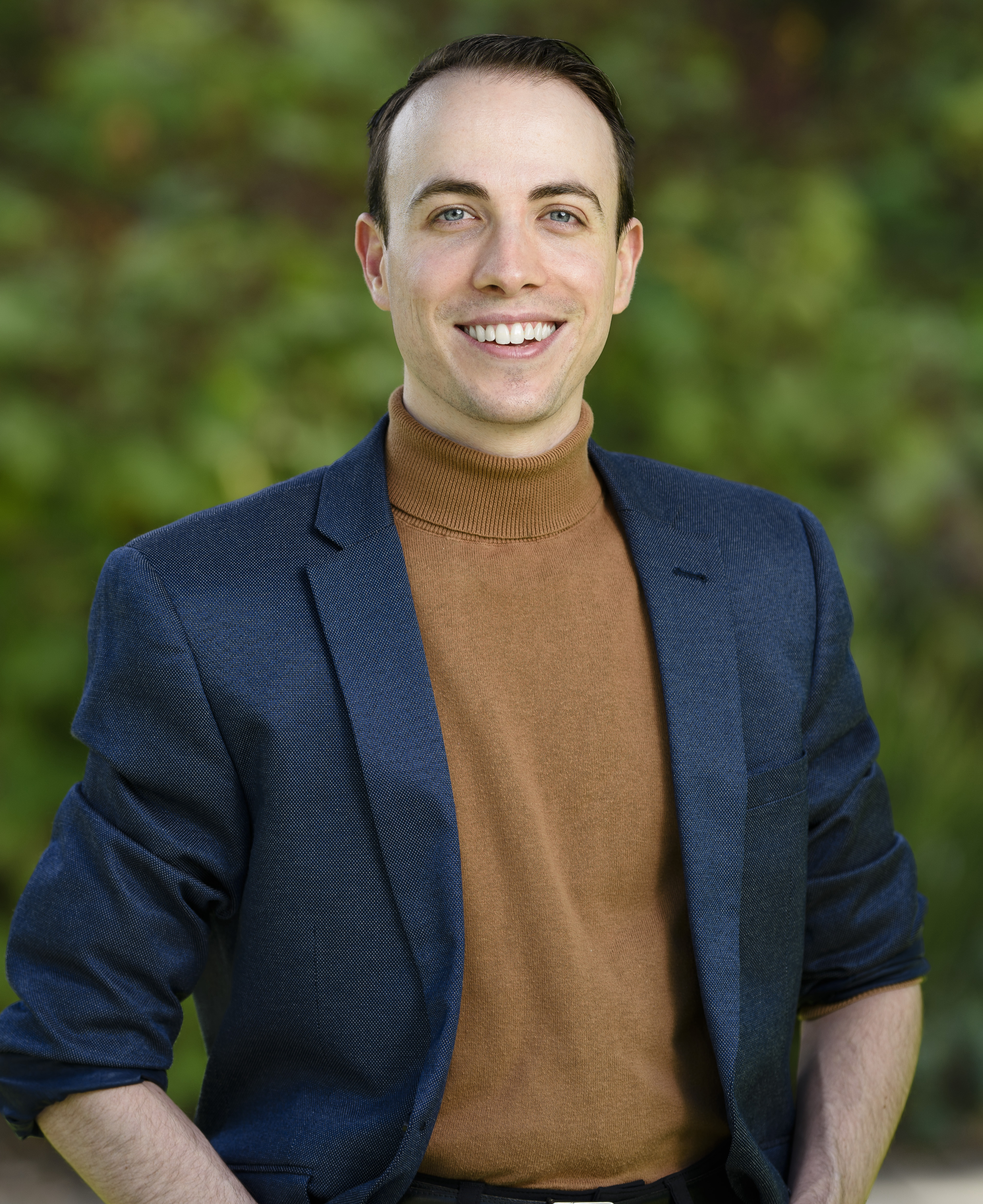 Headshot of Aiv Puglielli wearing a blue blazer, brown sweater, standing in front of blurred Green Foliage.