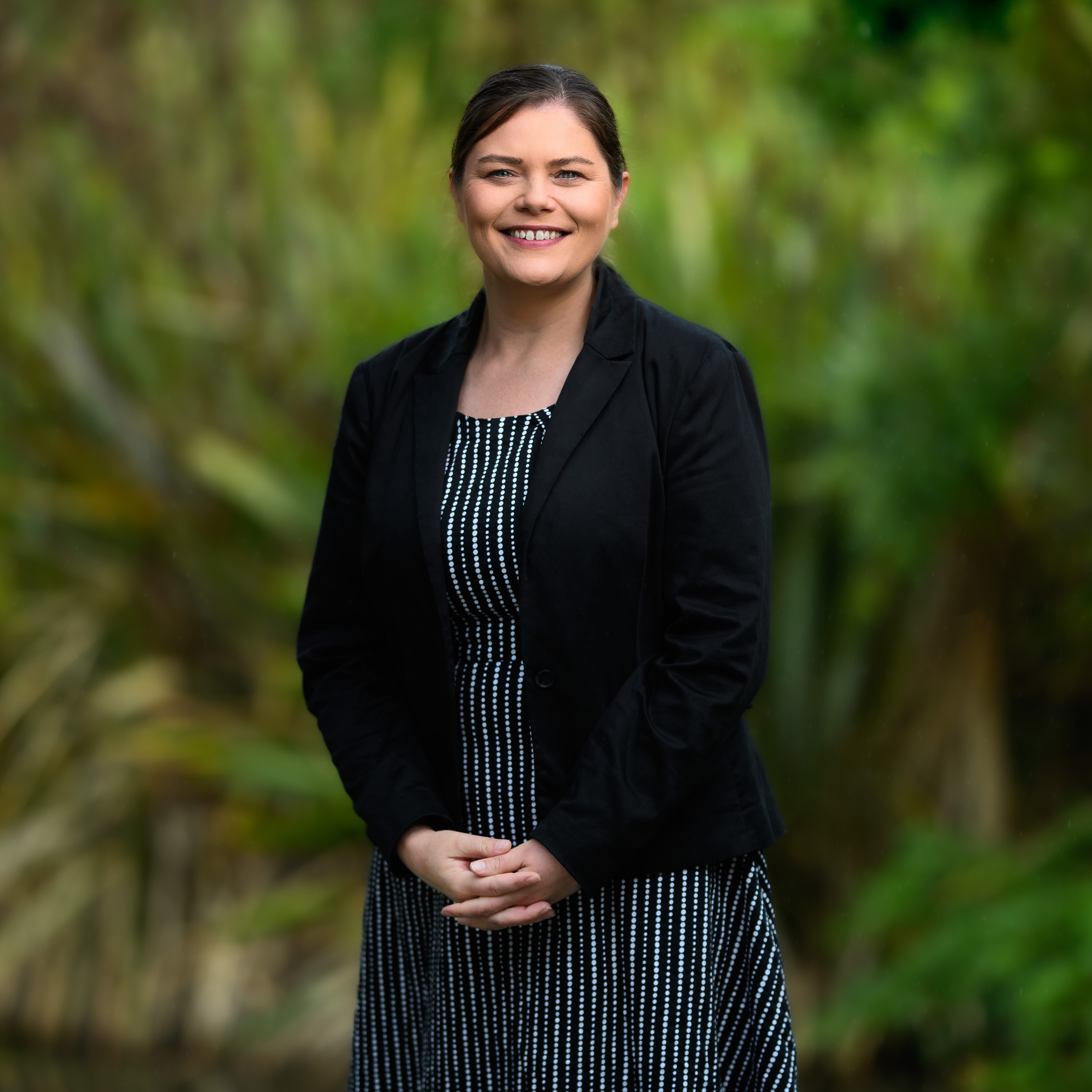 Jackie Carter, a woman with dark hair tied back, smiles warmly. She wears a black blazer and a patterned black-and-white dress, standing outdoors with a blurred green, leafy background.