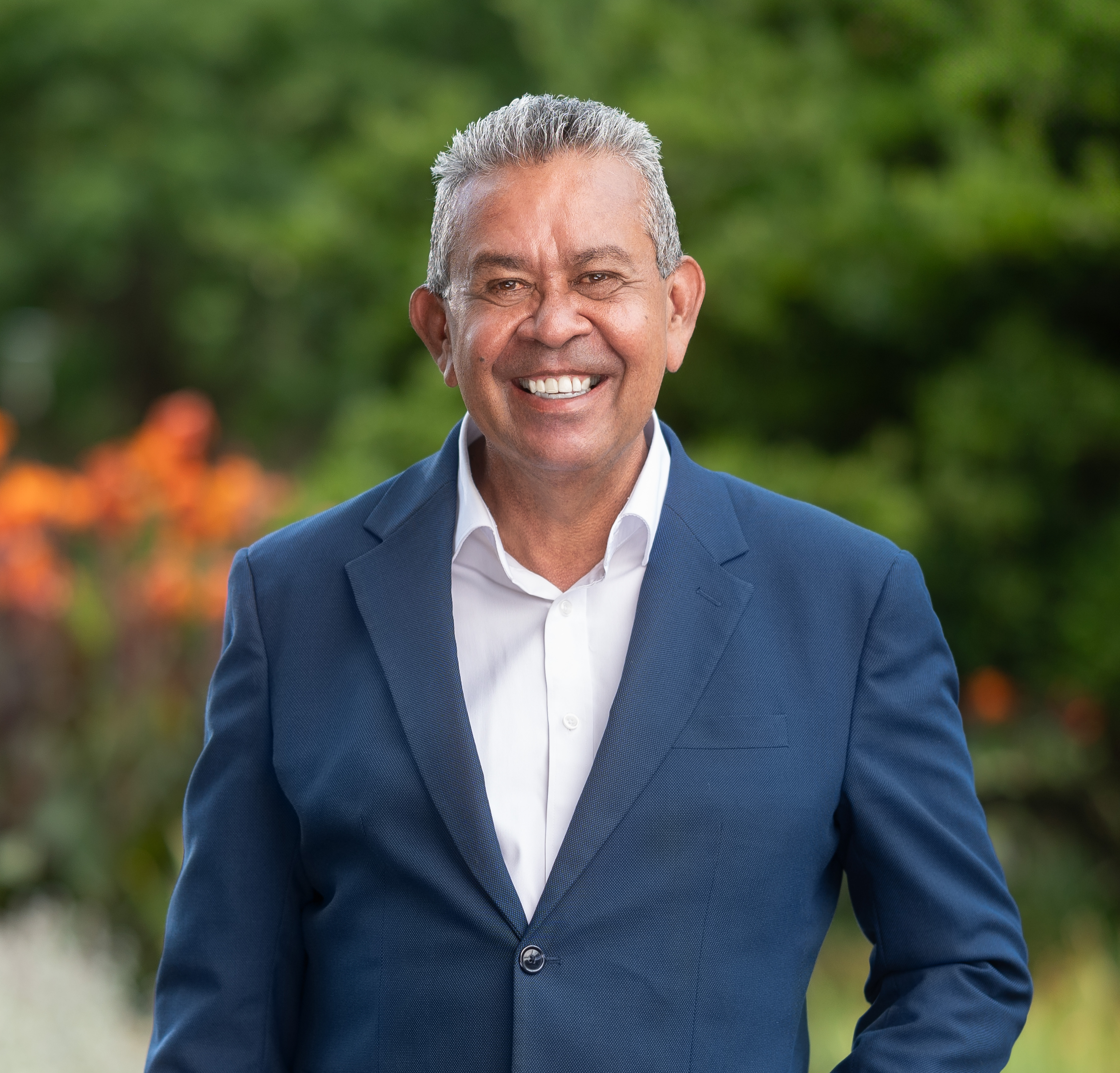 James Williams, an older man with short grey hair, smiles warmly while standing outdoors. He wears a navy blue blazer over a white shirt. His hand rests in his pocket, with a background of greenery and orange flowers.