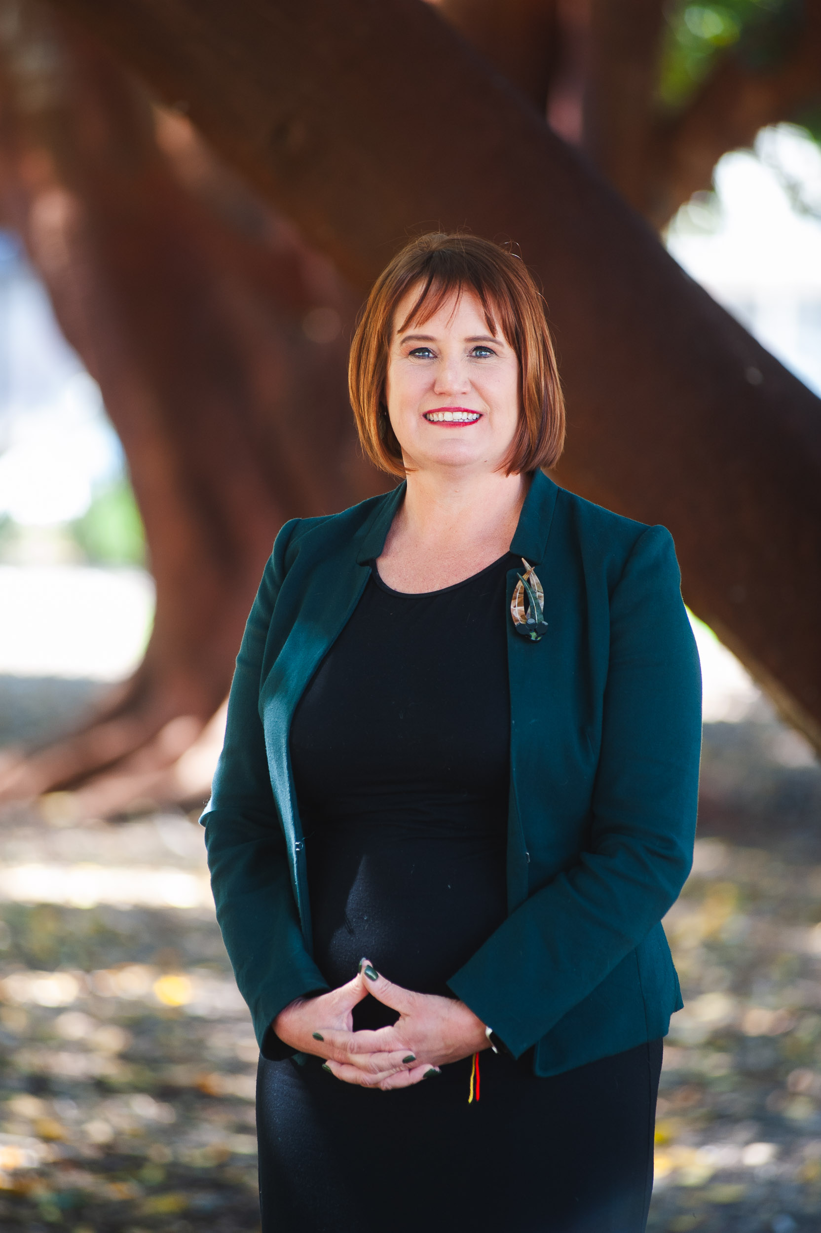 Janelle Sewell, a woman with brown hair wearing a black dress and green jacket, standing in a leafy park and smiling at the camera