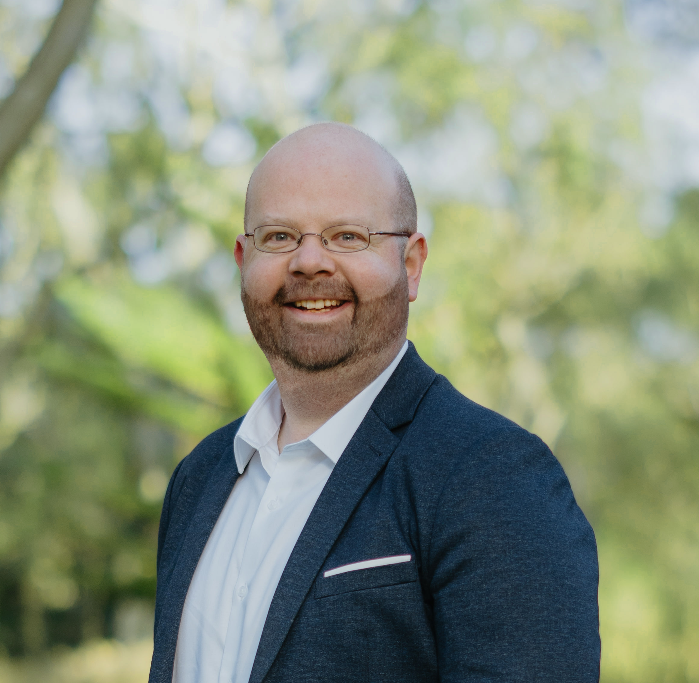 The image shows Jess Harper, a bald man with a beard and glasses, smiling outdoors. He is wearing a white shirt and a dark blue blazer, standing against a blurred green background of trees.