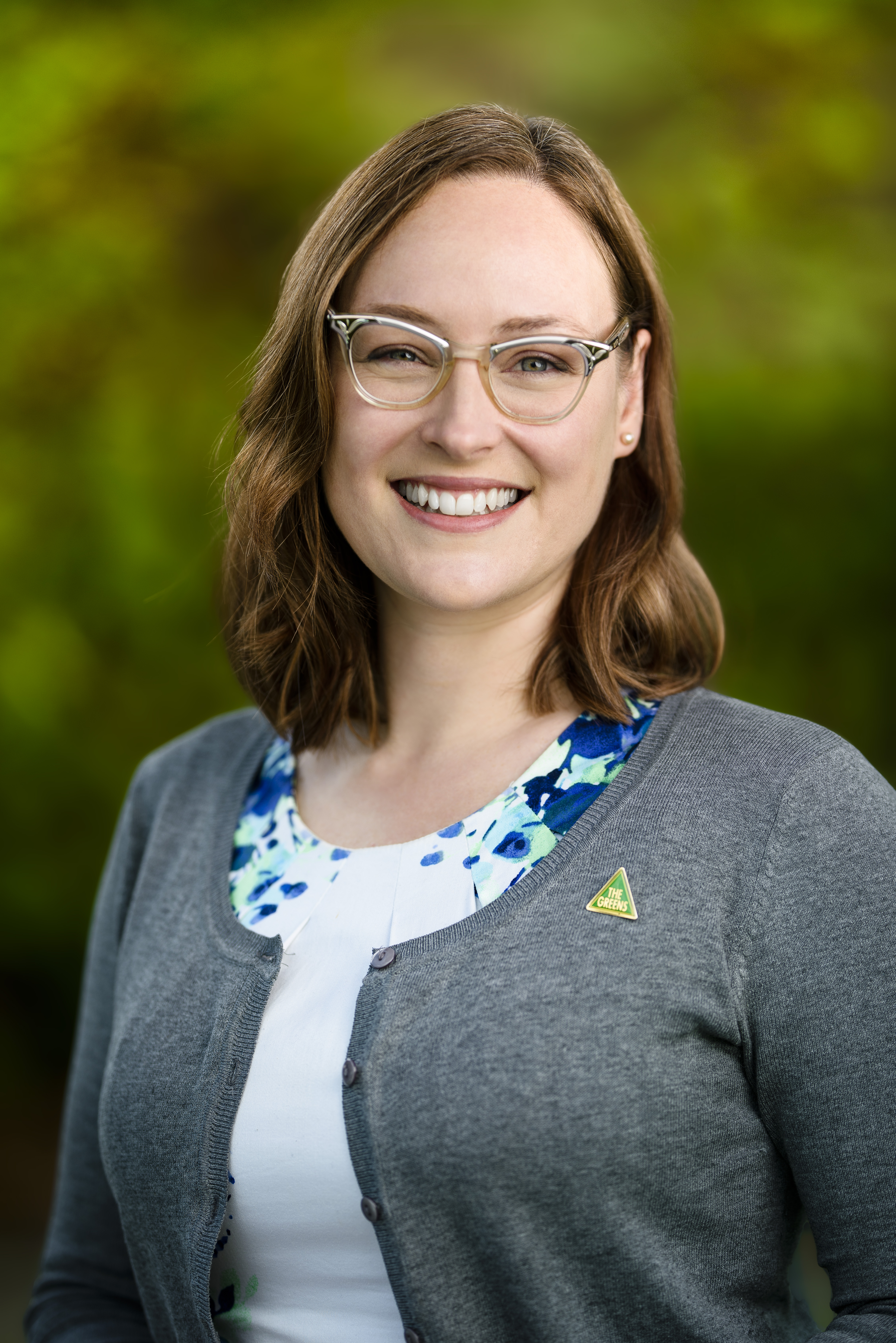 Headshot of Katherine Copsey wearing a white dress, grey cardigan, greens badge, glasses. Katherine is smiling and standing in front of blurred green foliage.