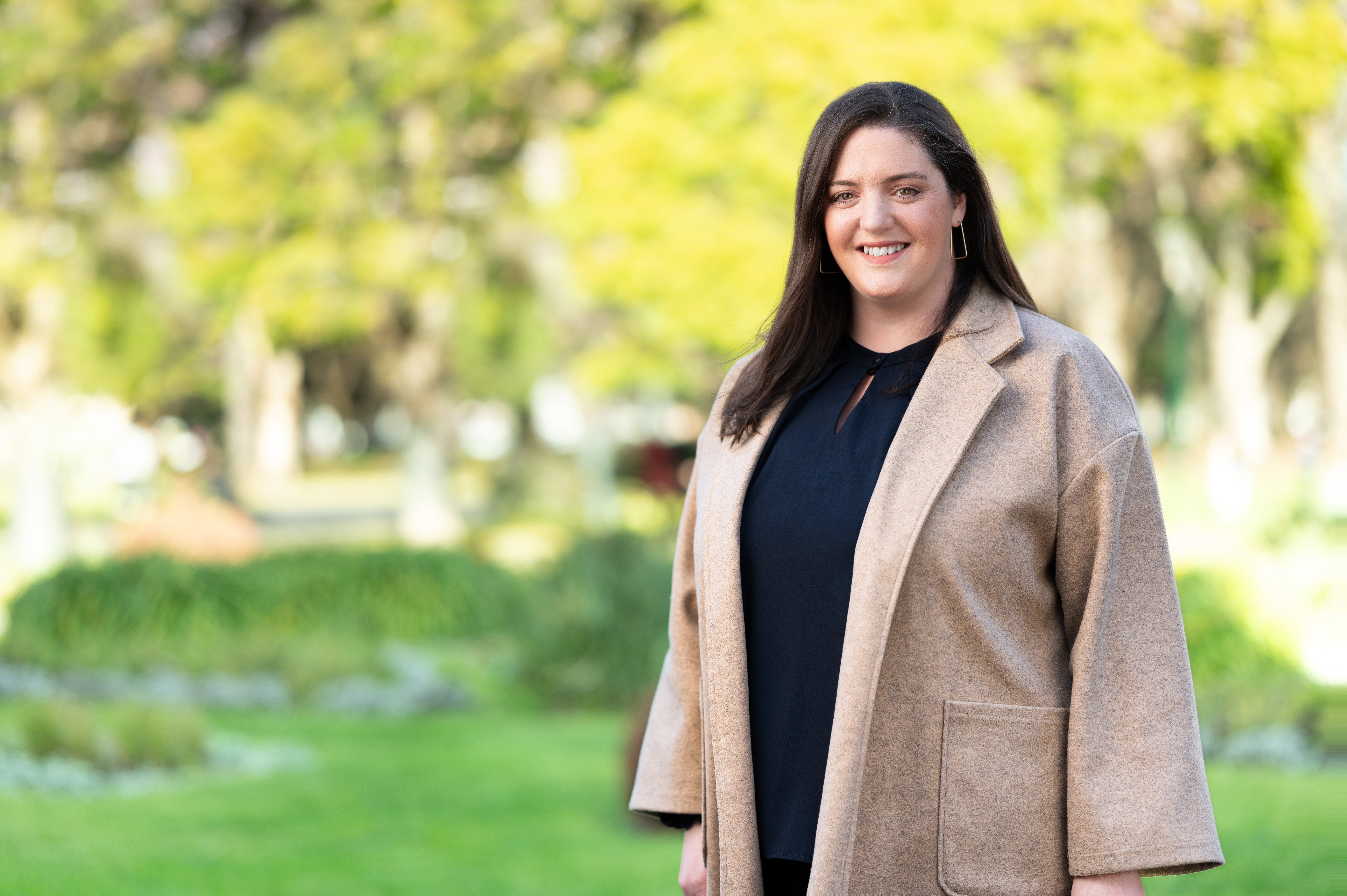 A smiling Kate McKay stands outdoors in a park, wearing a beige coat over a black top. The background is green and blurred.