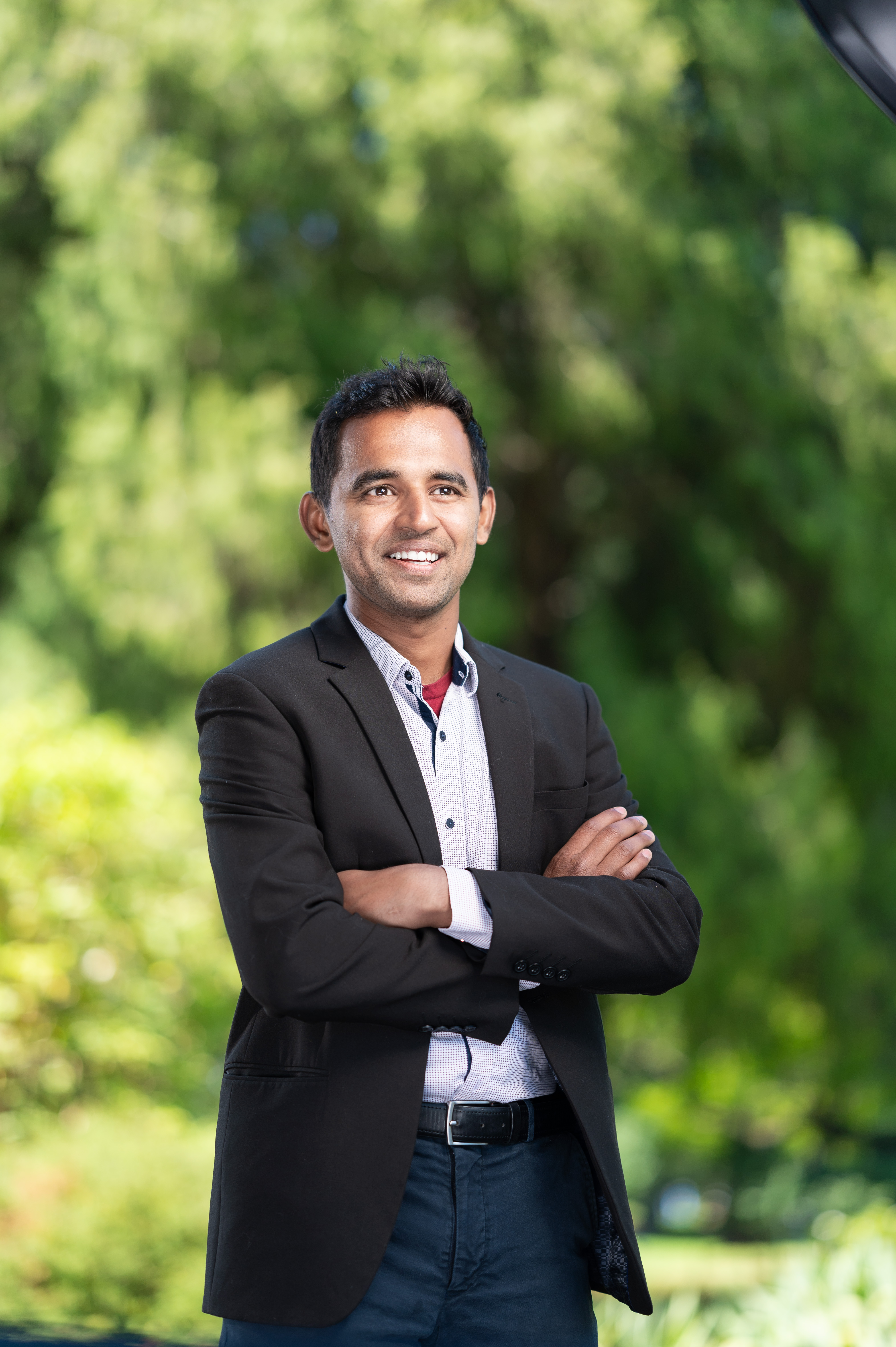 Lokesh Sangarya, a man with short black hair, smiles confidently with arms crossed. He wears a black blazer over a checkered shirt. The background is a lush green outdoor setting with sunlight filtering through the trees.