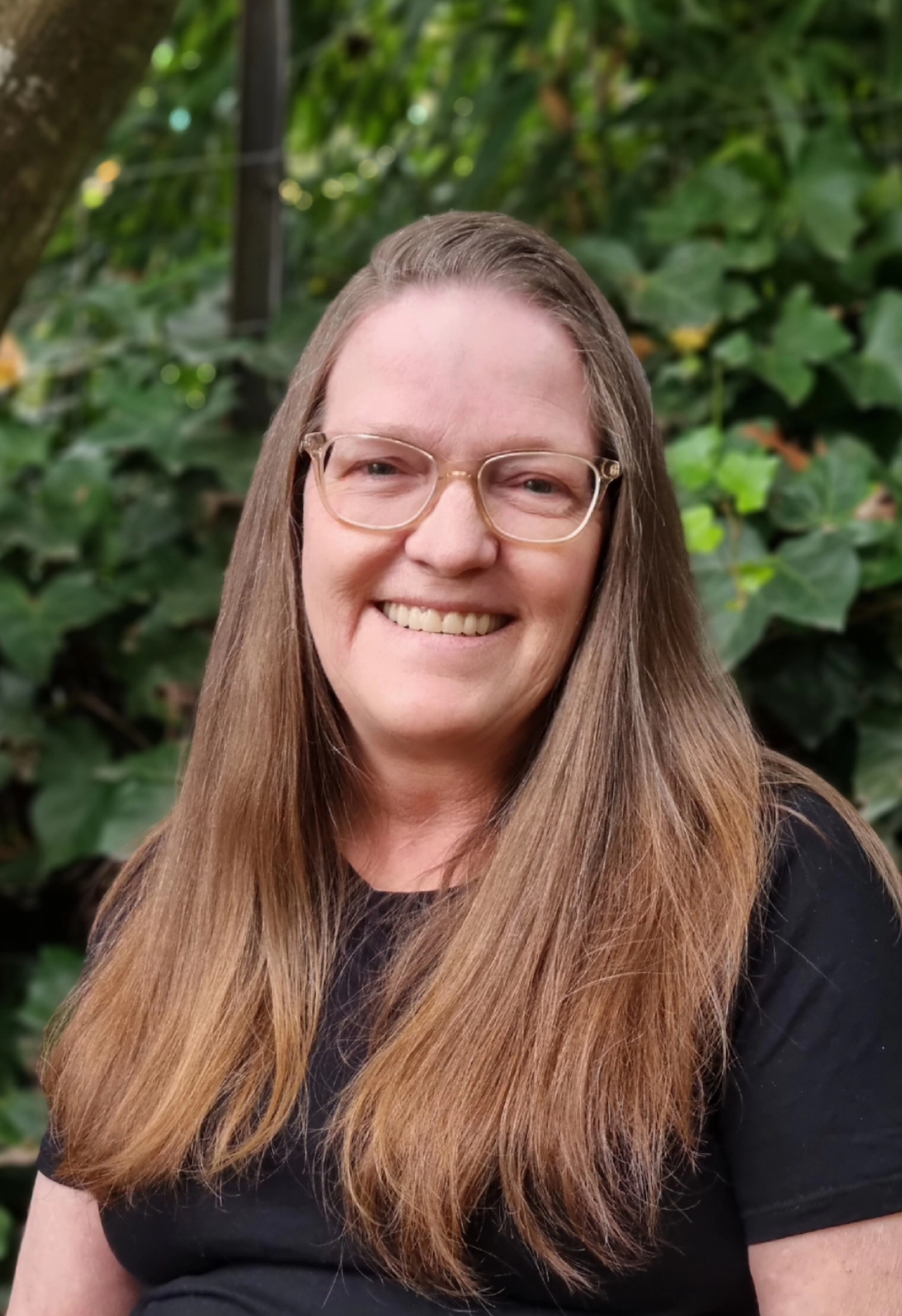 Lynda Wheelock in a black tshirt, wearing glasses and standing in front of greenery.