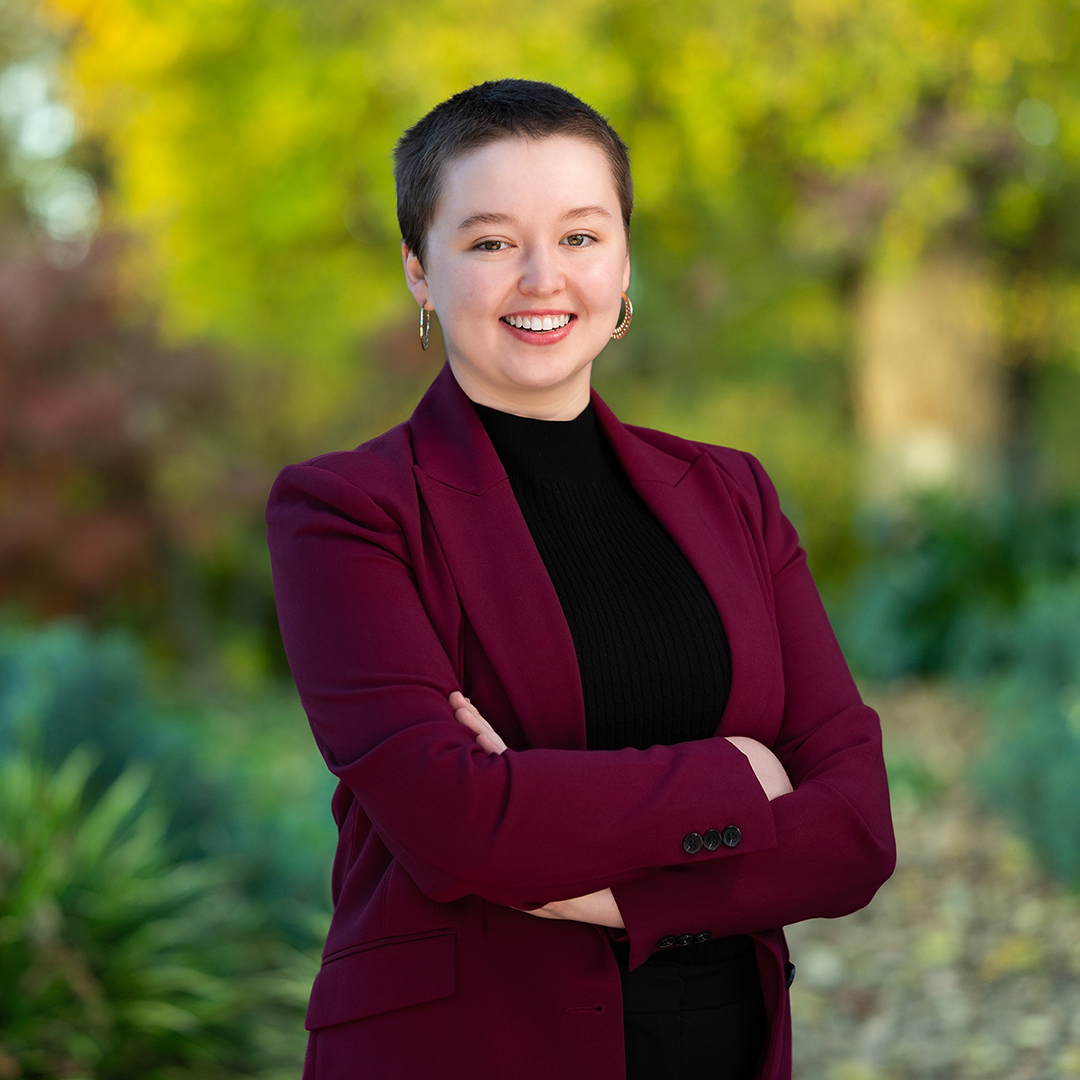 Marley, wearing a burgundy blazer over a black top, stands outdoors with a confident smile. The blurred greenery and autumn foliage in the background add a natural touch. Arms crossed, they appear both approachable and professional.