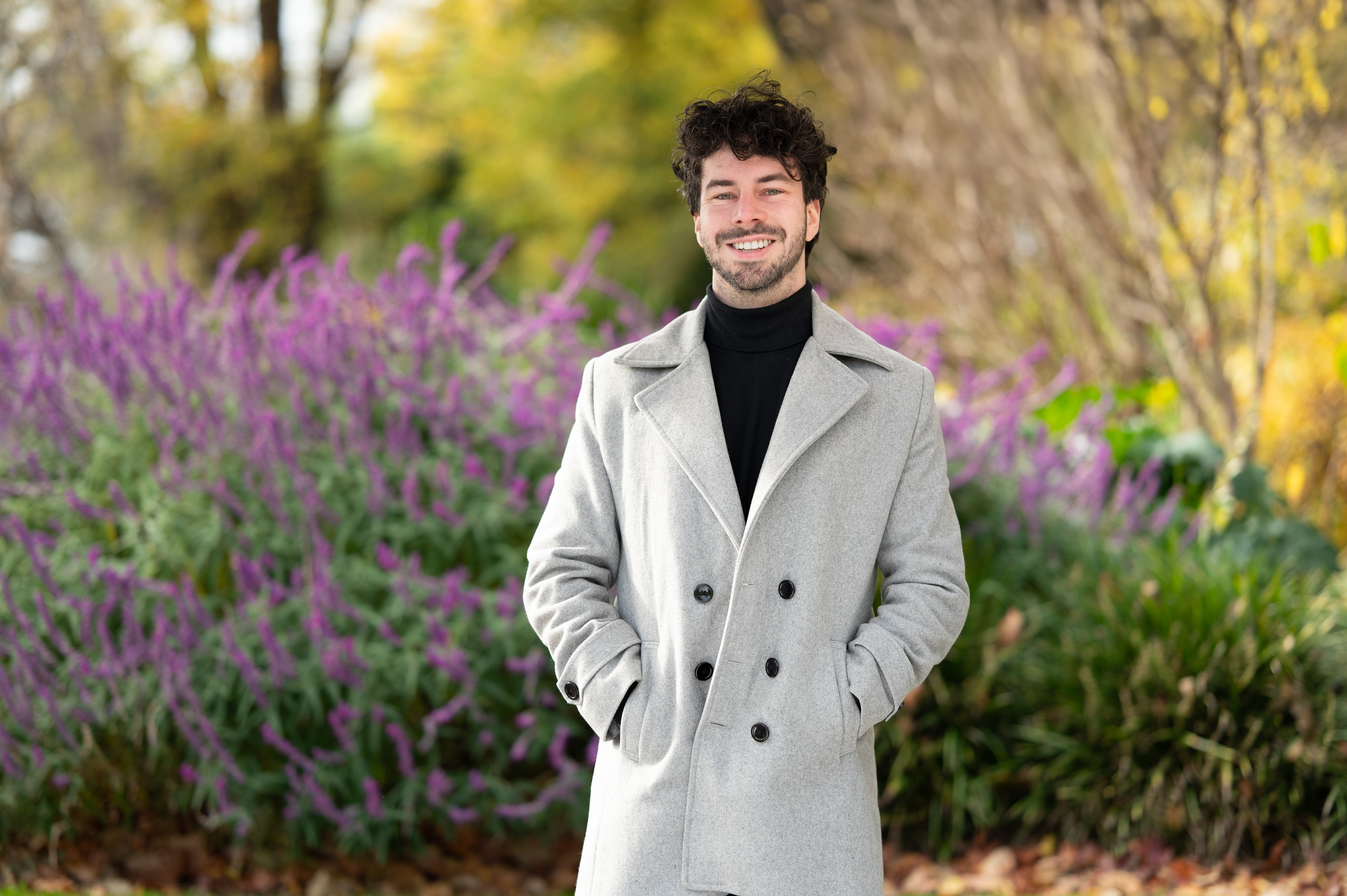 Mitchell Fuller stands outdoors in a light gray coat, smiling warmly. Behind him, lush green foliage and vibrant purple flowers create a colorful, serene background on an overcast day.