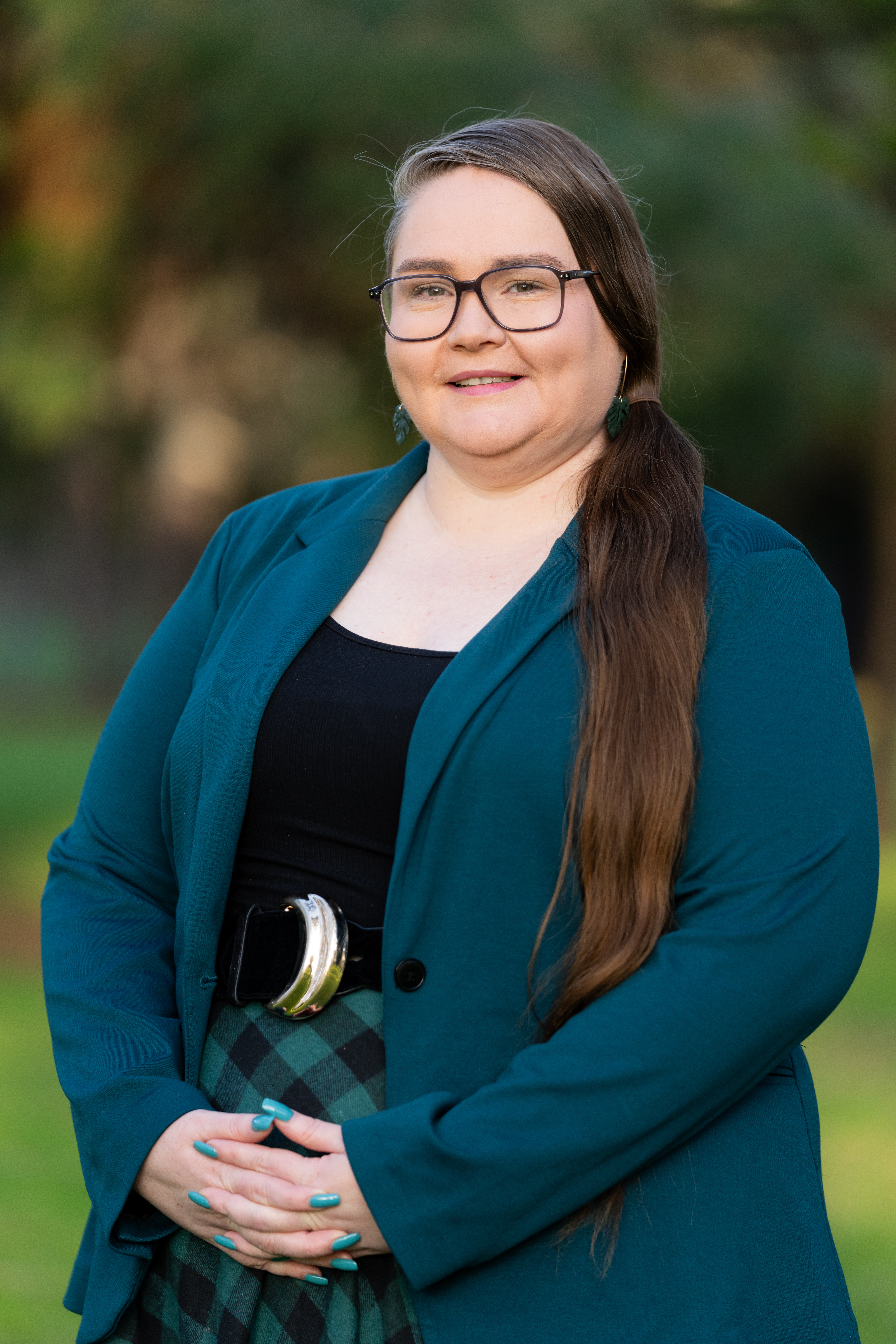 The image shows Theresa Slater, a woman with long brown hair pulled to the side, wearing glasses, a teal blazer, and a black top. She is standing outdoors against a blurred green background, smiling softly.