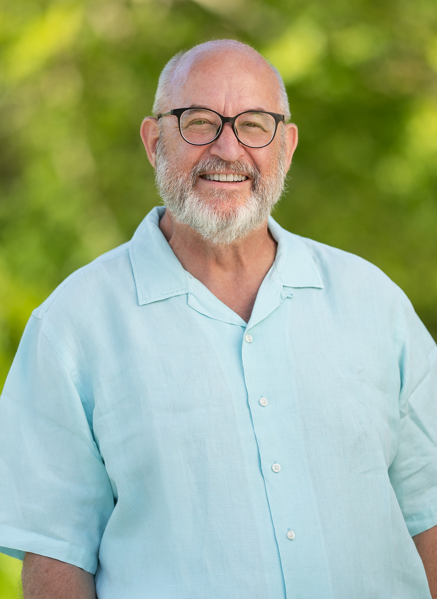 A man with a white beard, black glasses and blue button up shirt in front of a green background