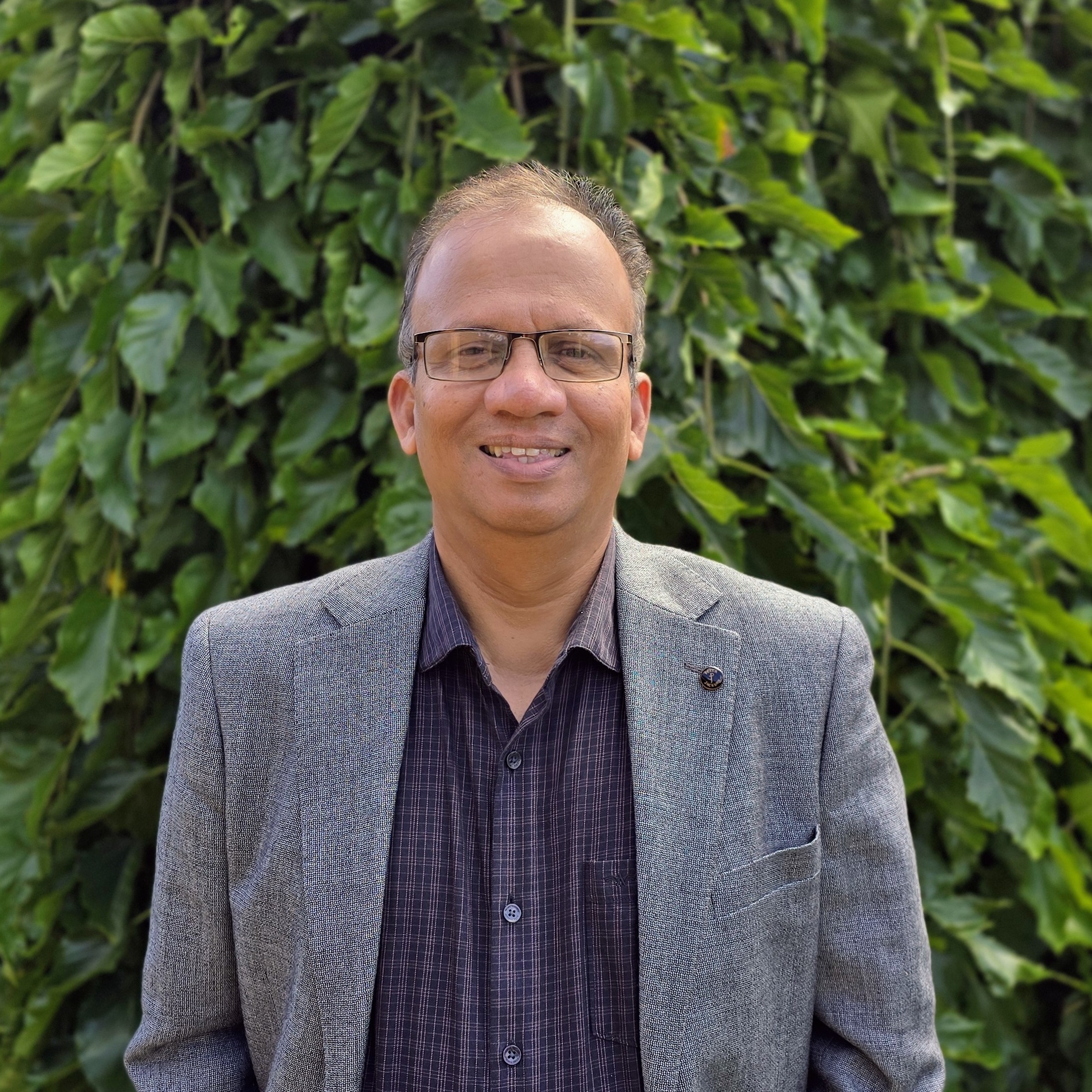 A man named Rifai, wearing glasses and a gray blazer over a dark checkered shirt, stands smiling against a backdrop of lush green foliage. The natural setting and his relaxed demeanor convey warmth and professionalism.