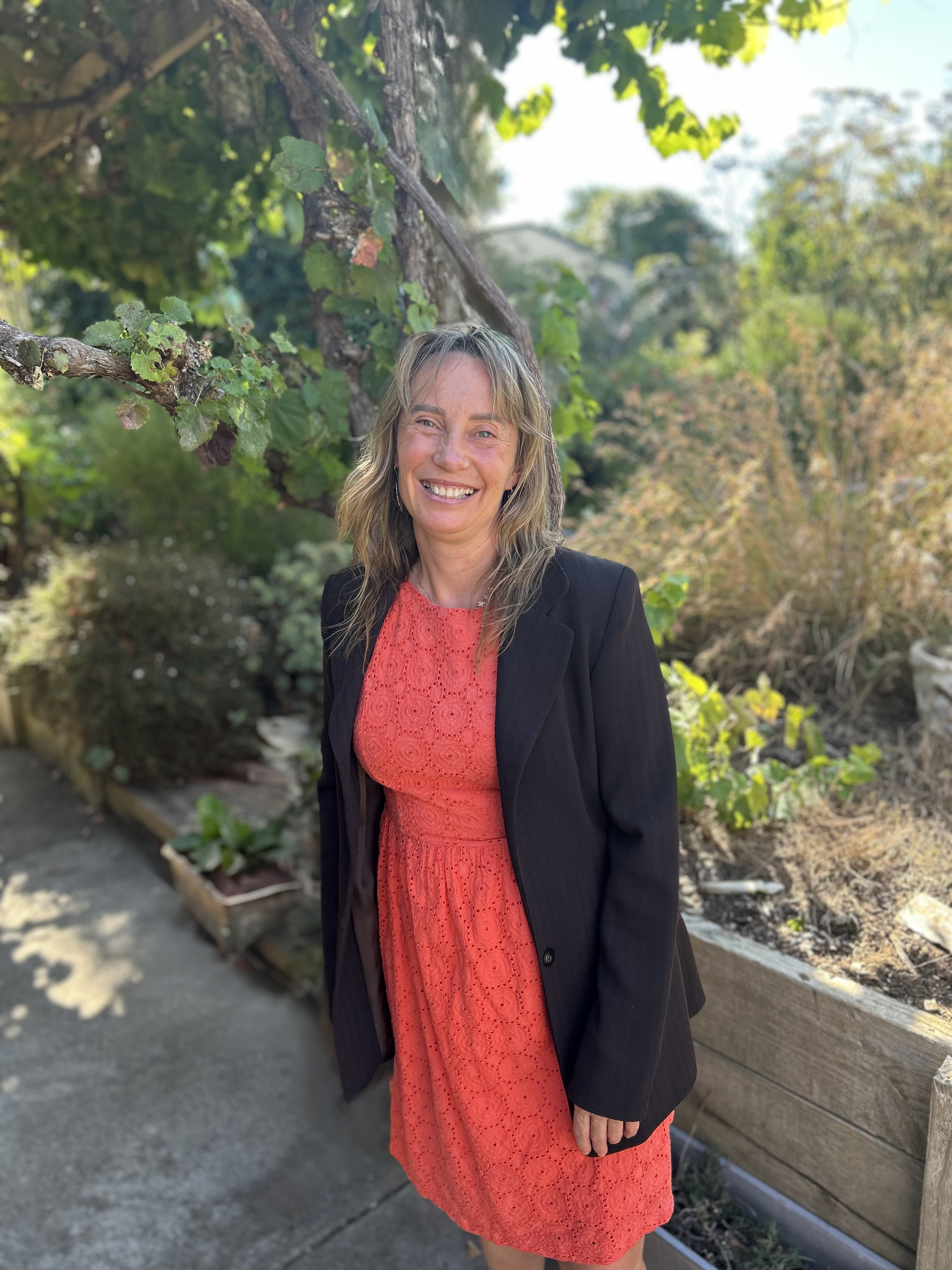 Rochelle Hine stands in a sunlit garden, smiling warmly. She wears a coral lace dress with a black blazer. Green vines and plants surround her, with a wooden planter and pathway in the background.