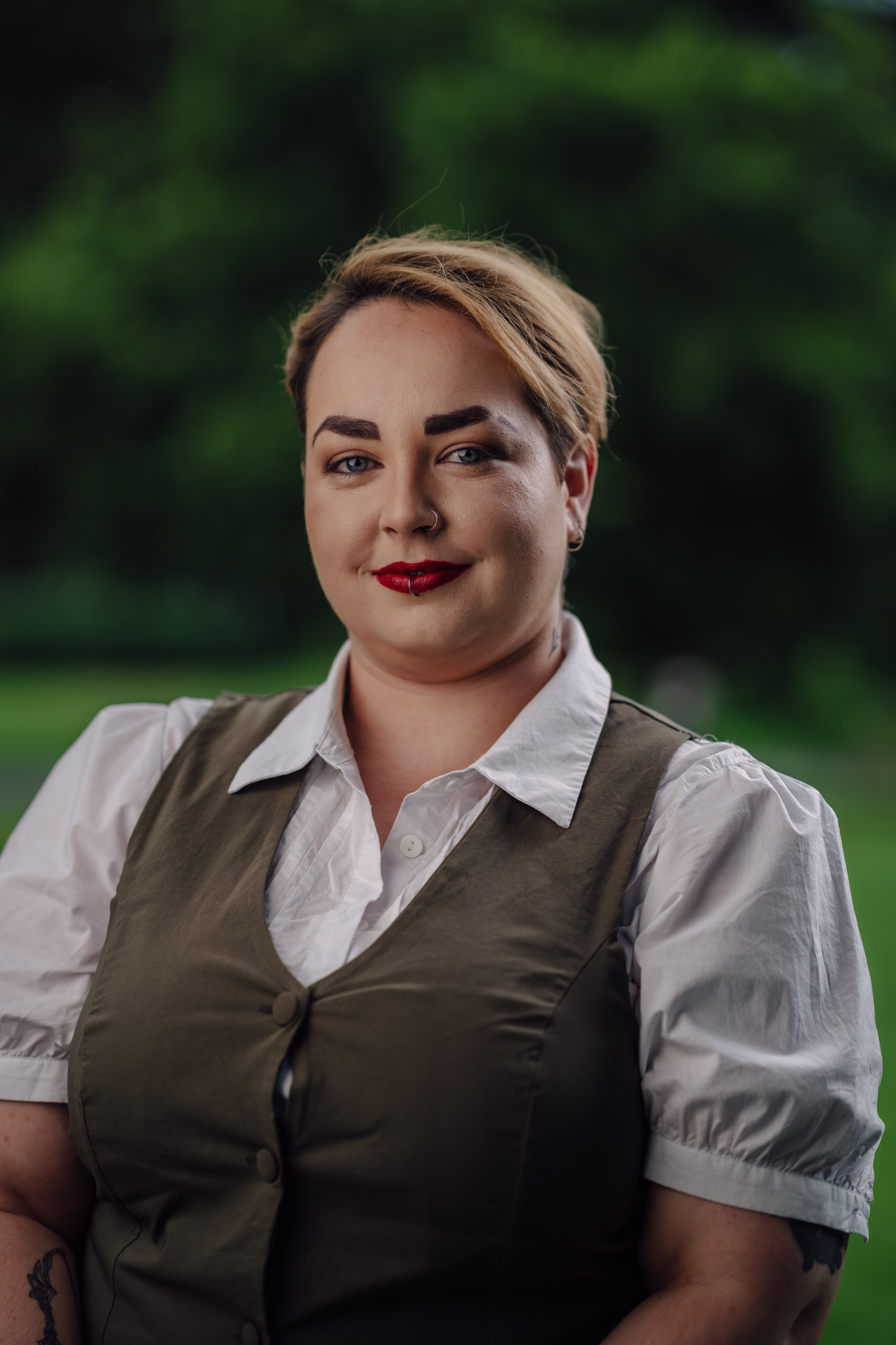 Sarah Newman, a woman with short blonde hair and red lipstick, smiles softly. She wears a white shirt and olive vest, sitting outdoors with a blurred green background.