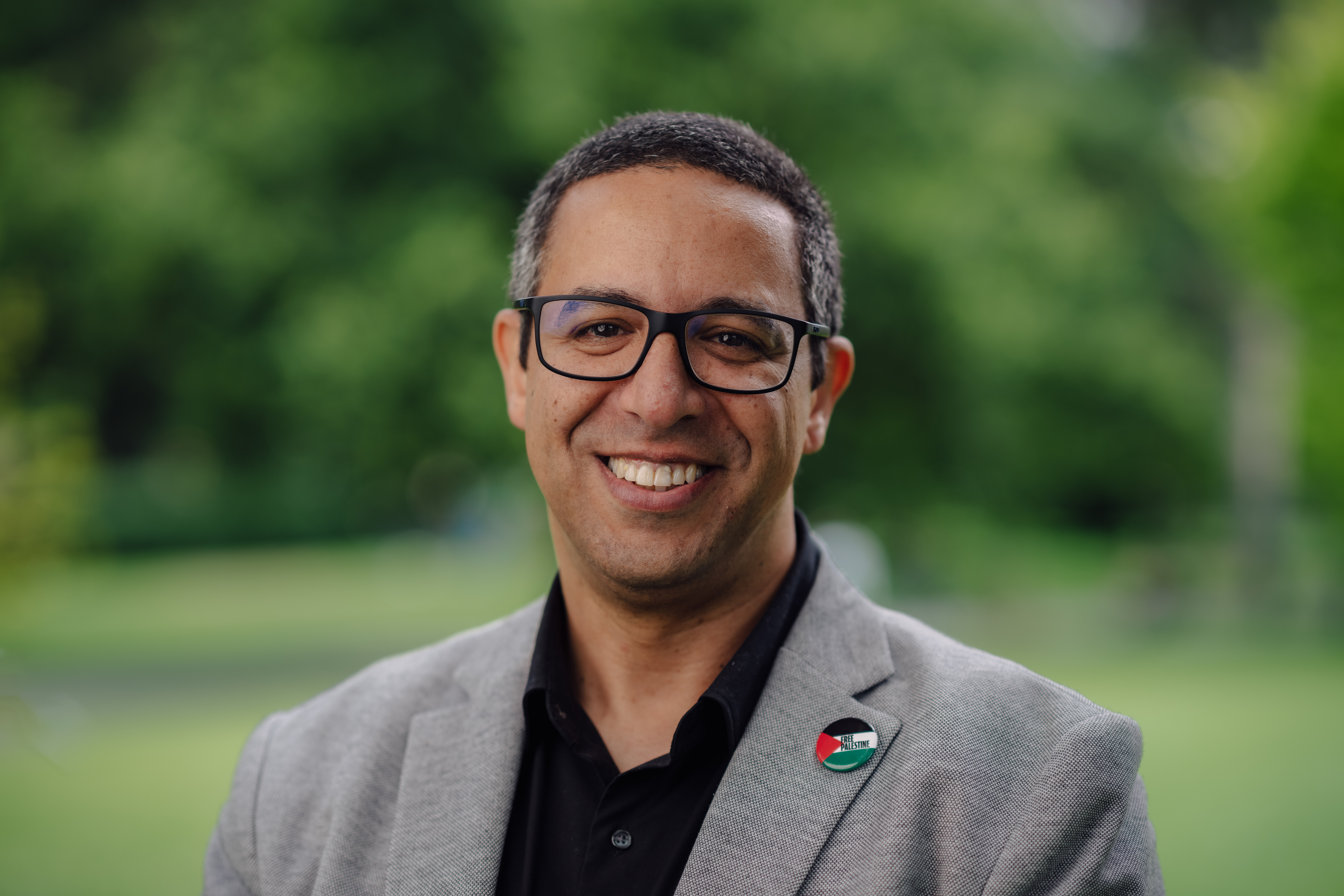  Mohamed El-Mastri, wearing glasses, a light gray blazer, and a black shirt, smiles warmly. A badge with the Palestinian flag is pinned to his blazer. He stands outdoors with a blurred green background.