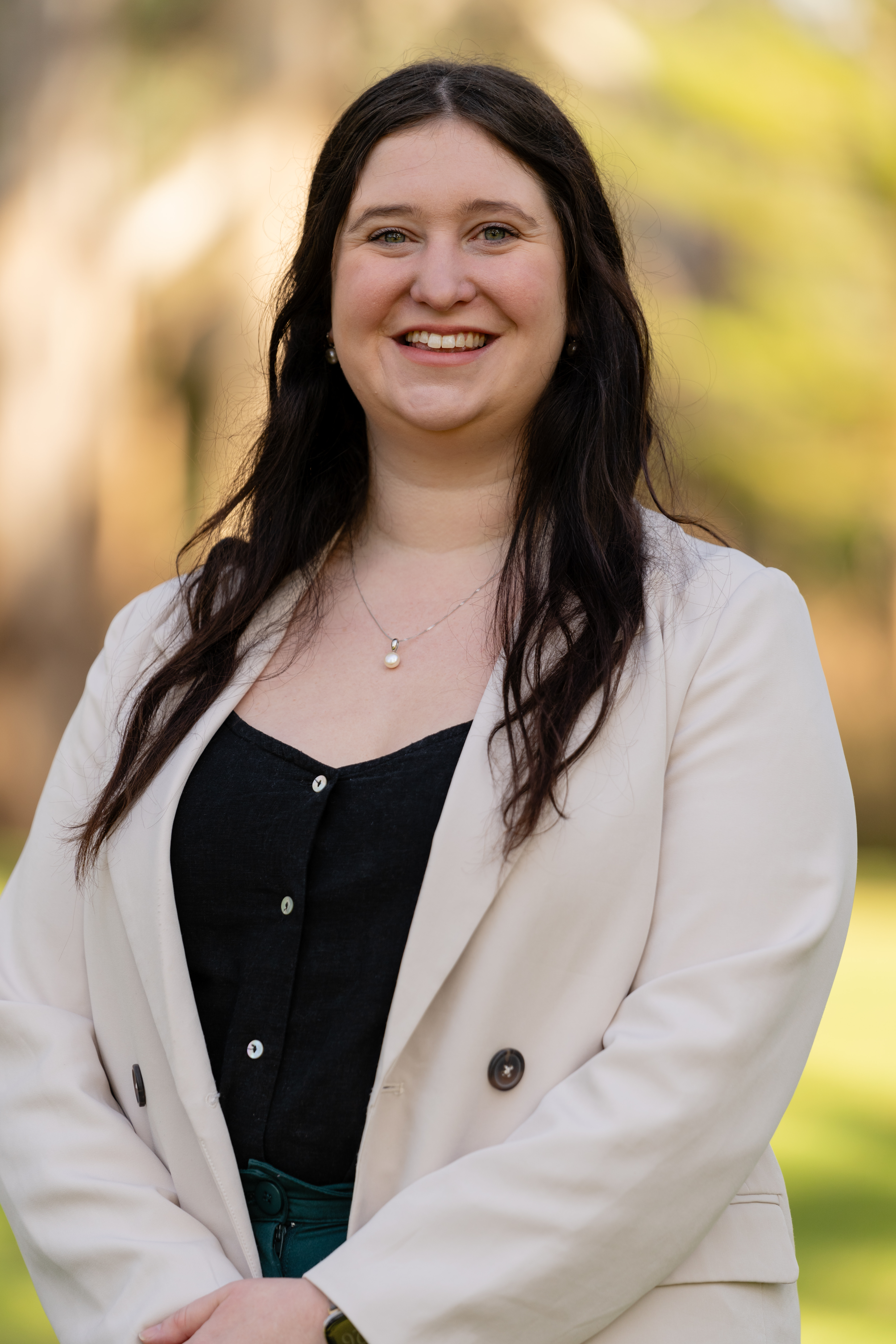 Sian is standing outdoors, wearing a beige blazer over a black top. She is smiling warmly, with long, dark hair. The background is softly blurred, featuring green and brown tones from trees and foliage.