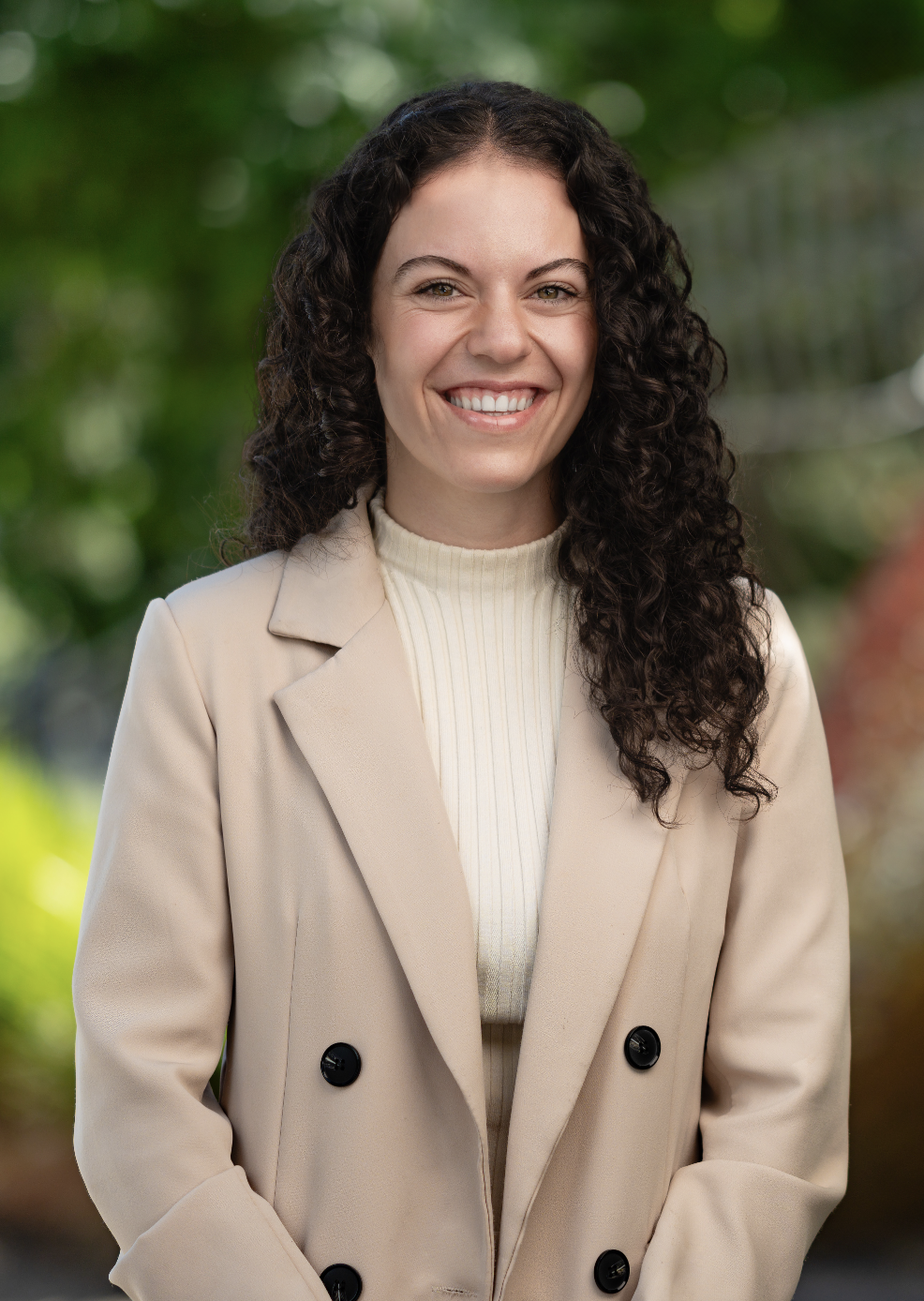 Angelica smiling at the camera in a beige jacket and cream top with long black curly hair