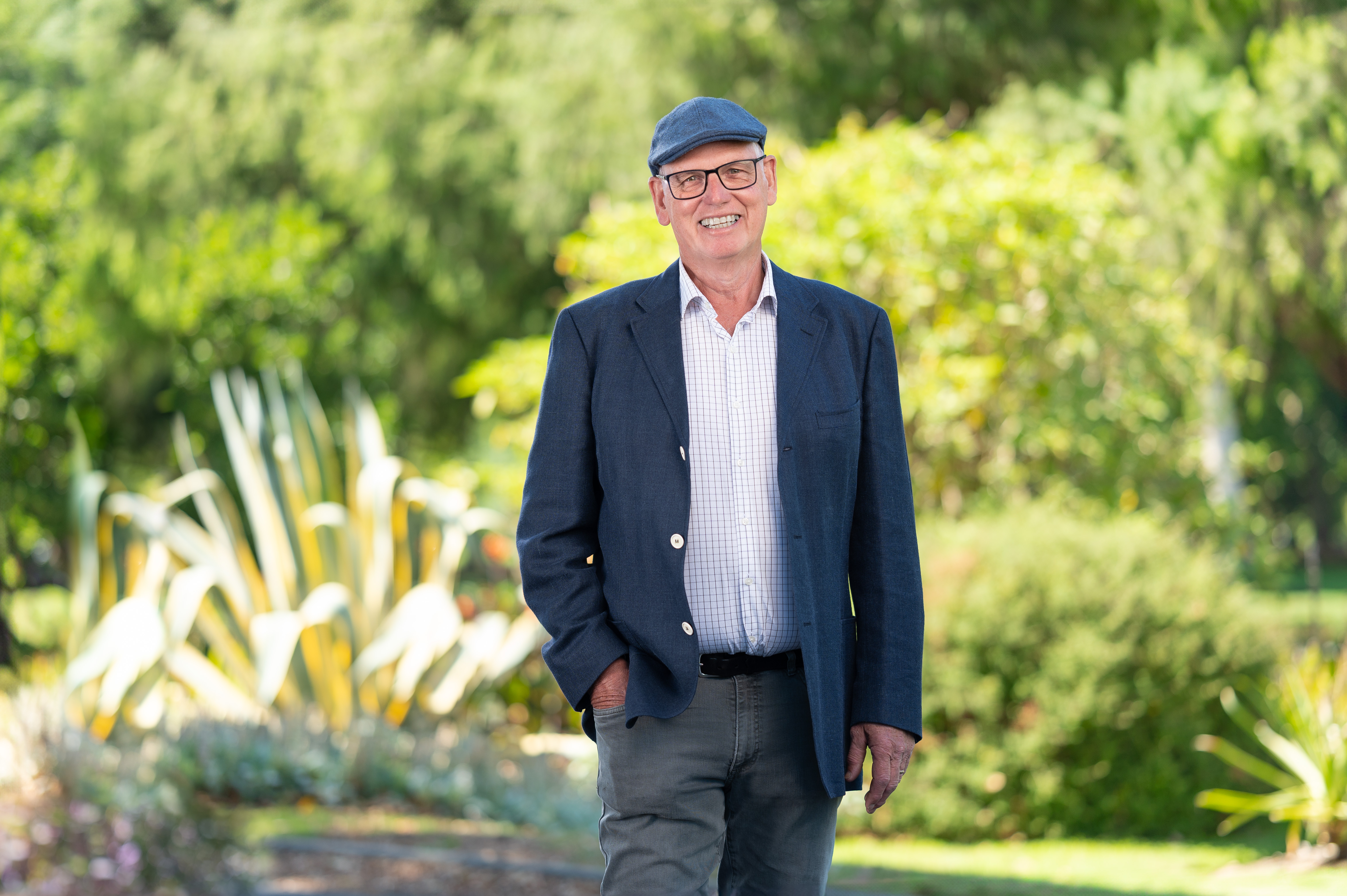 Terence Steele stands outdoors, smiling warmly. He wears a navy blazer, checkered shirt, gray pants, and a flat cap. Lush greenery and large variegated plants surround him in a sunlit park setting.