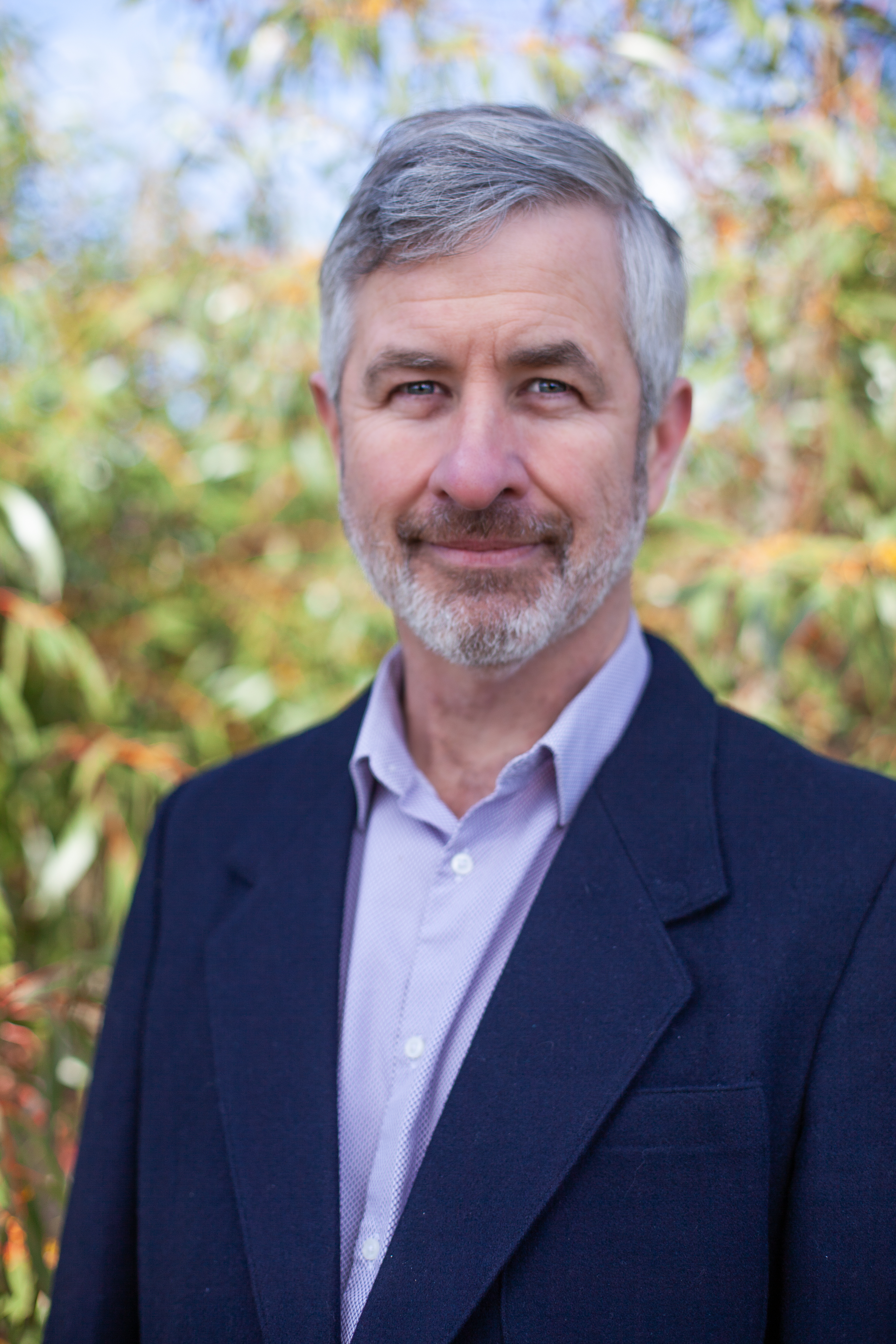  A middle-aged man with gray hair and a beard, named Tim Drylie, is wearing a dark blazer and light shirt. He is standing outdoors with blurred greenery in the background.