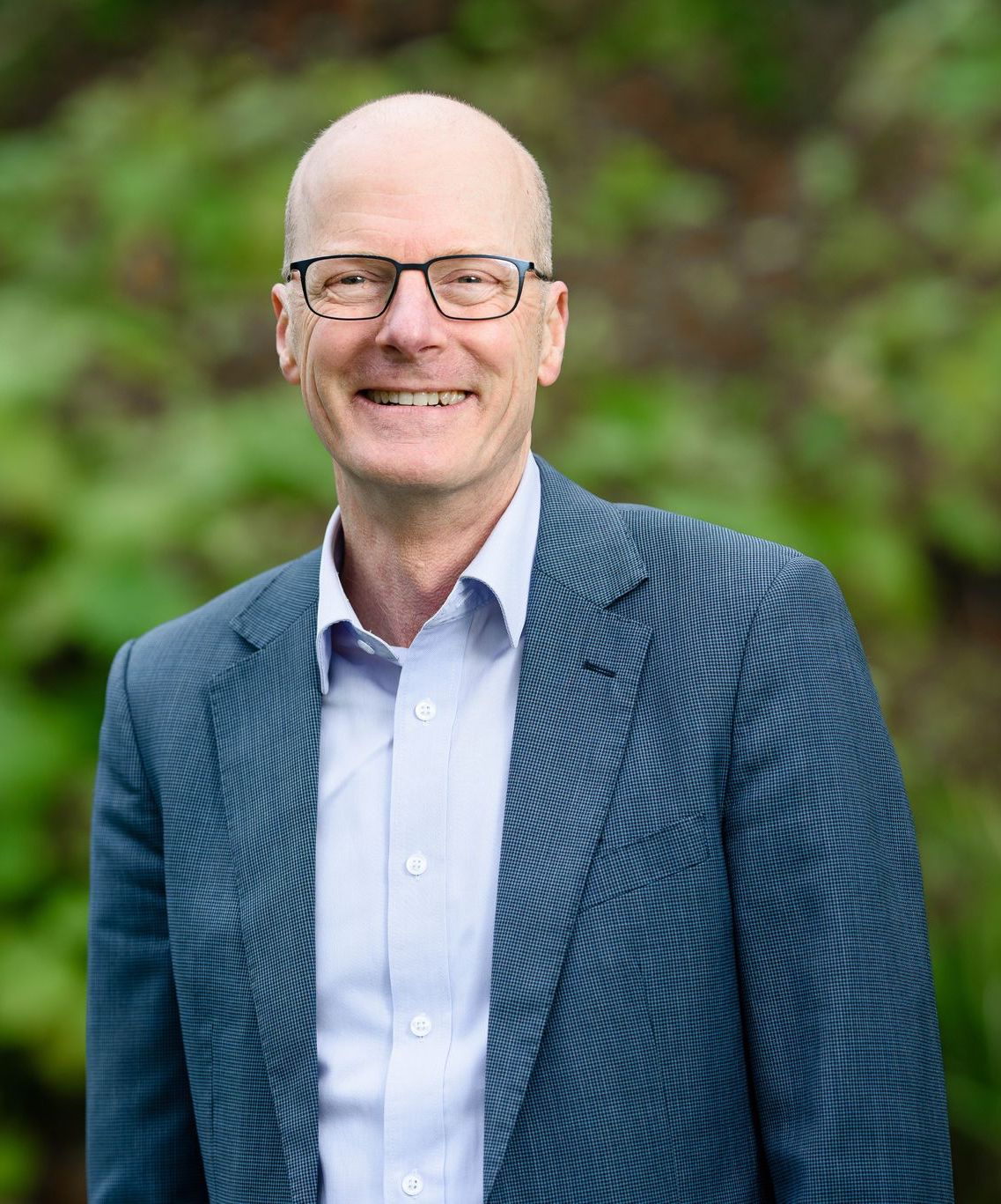 Tim is smiling whilst wearing glasses, a jacket and shirt and standing in front of blurred green foliage.