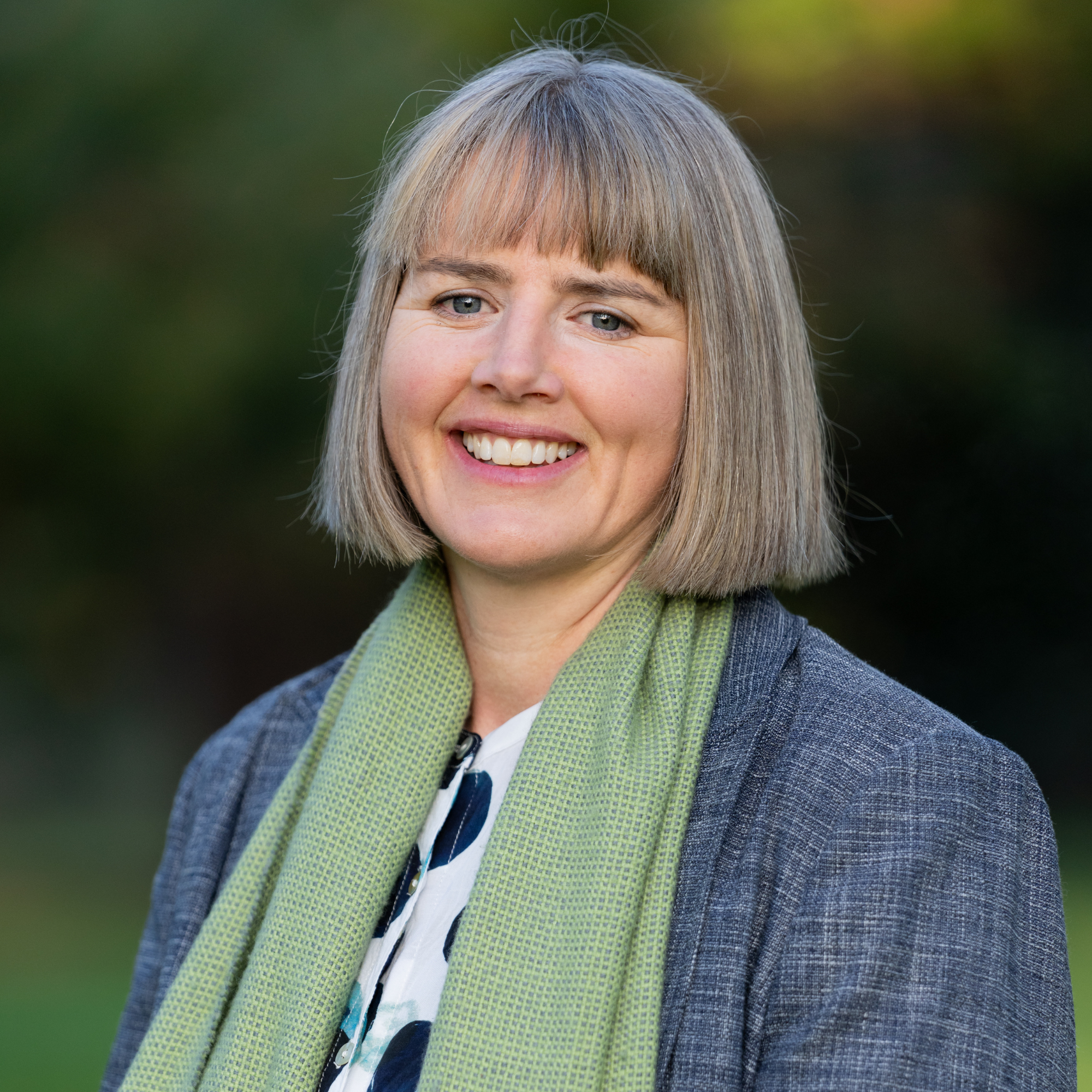 The image shows Izzy Scherrer, a woman with straight, shoulder-length gray hair and bangs, smiling outdoors. She is wearing a gray blazer, a green scarf, and a patterned top, against a blurred background of greenery.