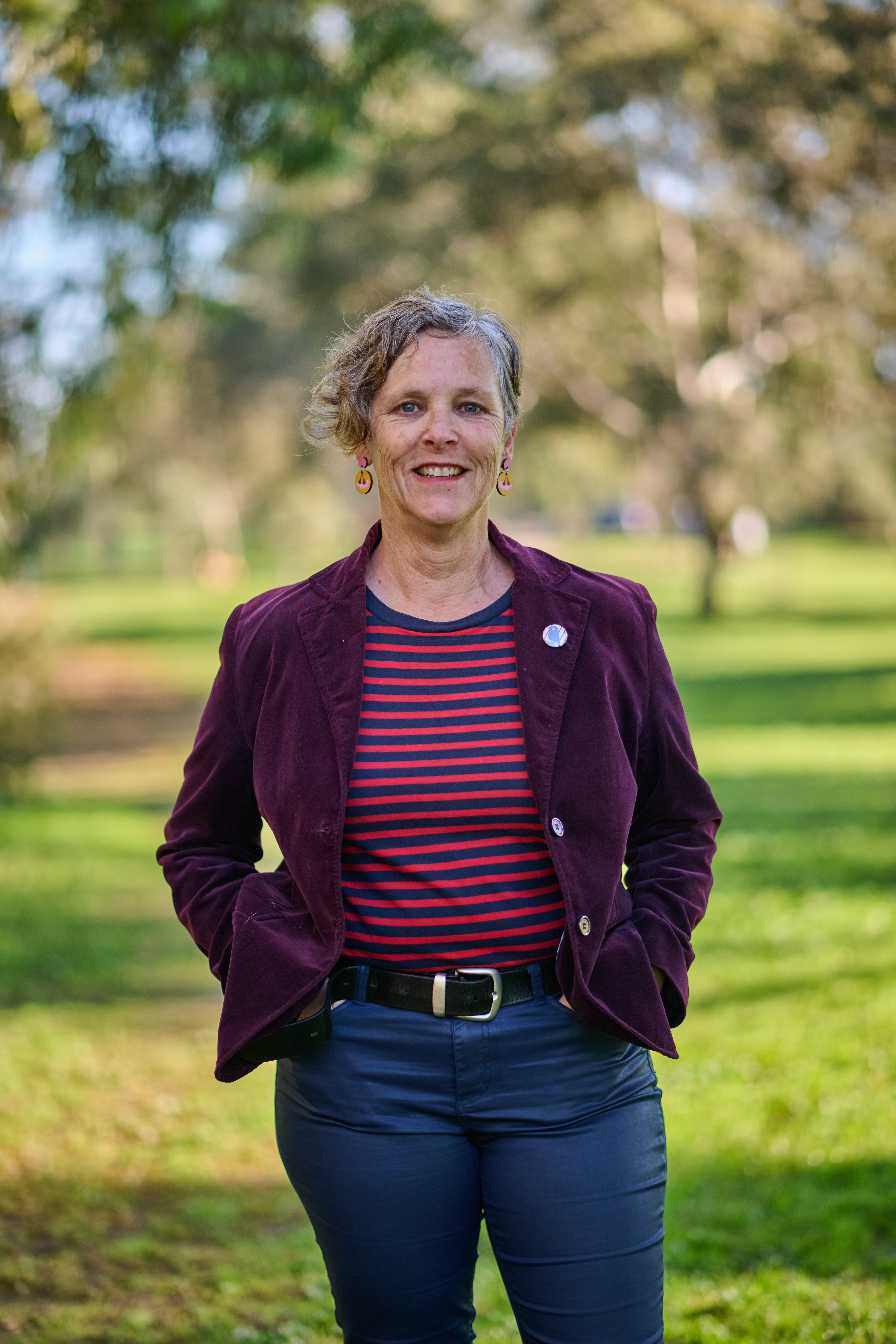 A woman with short brown hair, a purple blazer, stripy red and blue shirt and blue pants. Standing in a park with hands in pockets.