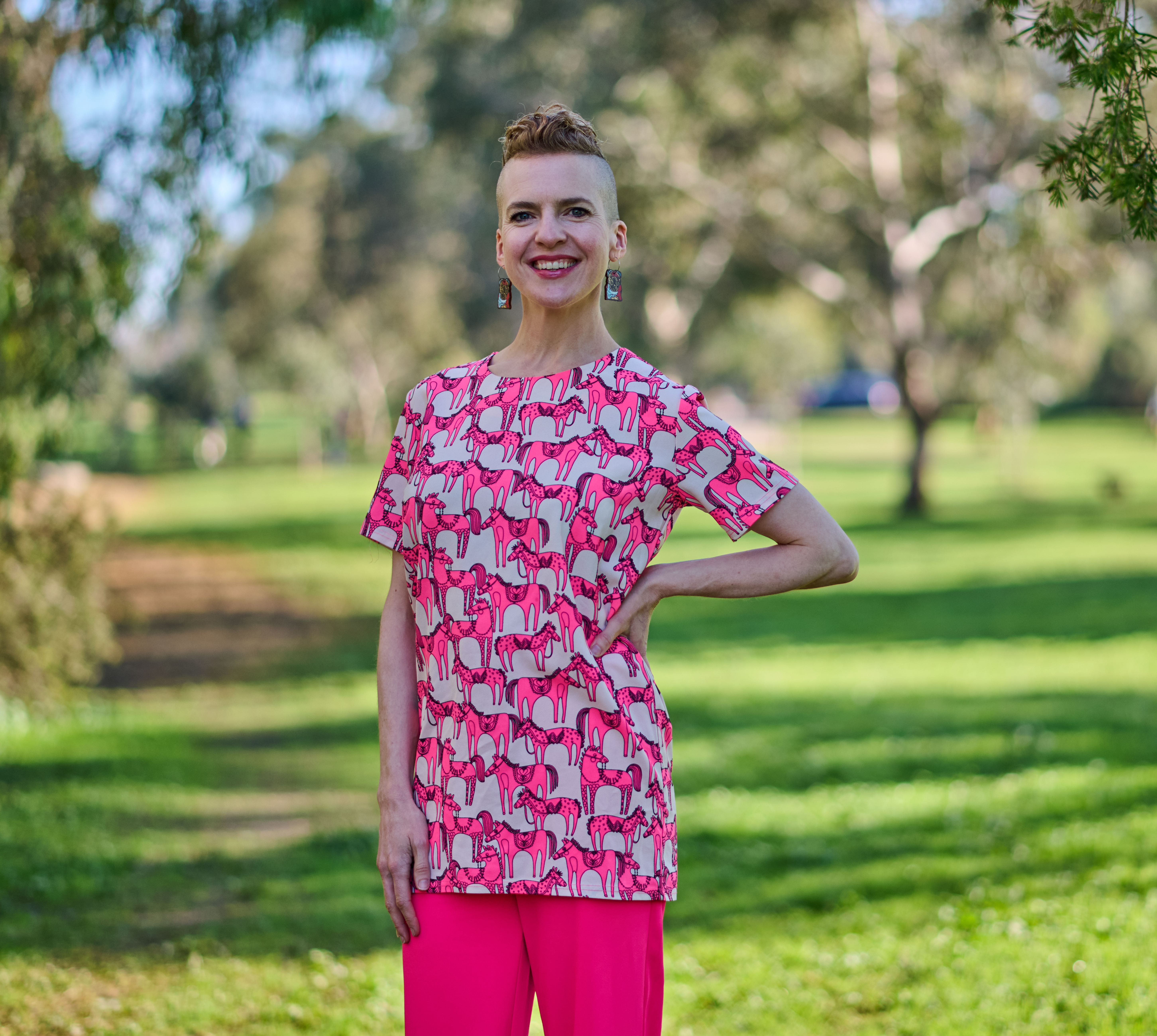 A person with short hair, wearing a pink top and pink pants standing with one hand on their hip in a green park smiling.